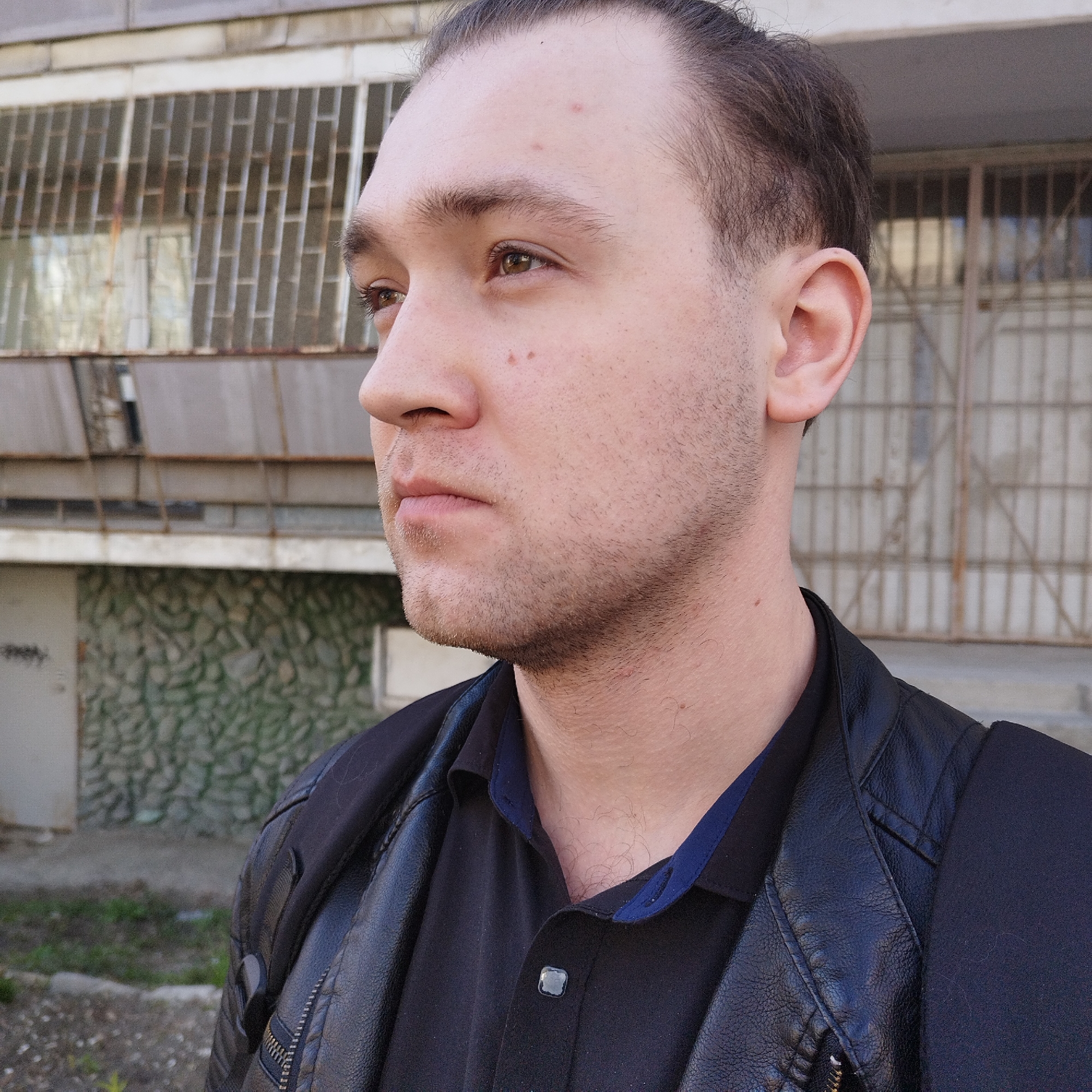 A young man is captured in a profile portrait, facing towards the viewer's right, set against an urban background. He appears to be in his twenties or early thirties, with dark, neatly styled hair and short facial stubble. His brown eyes are open, and he maintains a neutral, contemplative expression as he looks off into the distance. Several small moles are visible on his face and forehead.
He is wearing a black polo shirt with a dark blue trim on the collar, underneath a black, leather-like jacket. A black strap, likely from a backpack, is visible over his left shoulder.
The background consists of an old, somewhat worn building, typical of an urban environment. It features a facade with gridded windows, some of which appear to have metal bars or frames. The lower section of the building wall is covered in a distinctive green and gray textured finish, possibly stone or mosaic. The ground immediately behind him is bare earth with sparse patches of grass and weeds. The lighting suggests it is daytime, possibly overcast, creating soft, even illumination. The overall scene reflects an everyday urban setting, consistent with locations in Yekaterinburg, Russia. No discernible text is visible in the image.