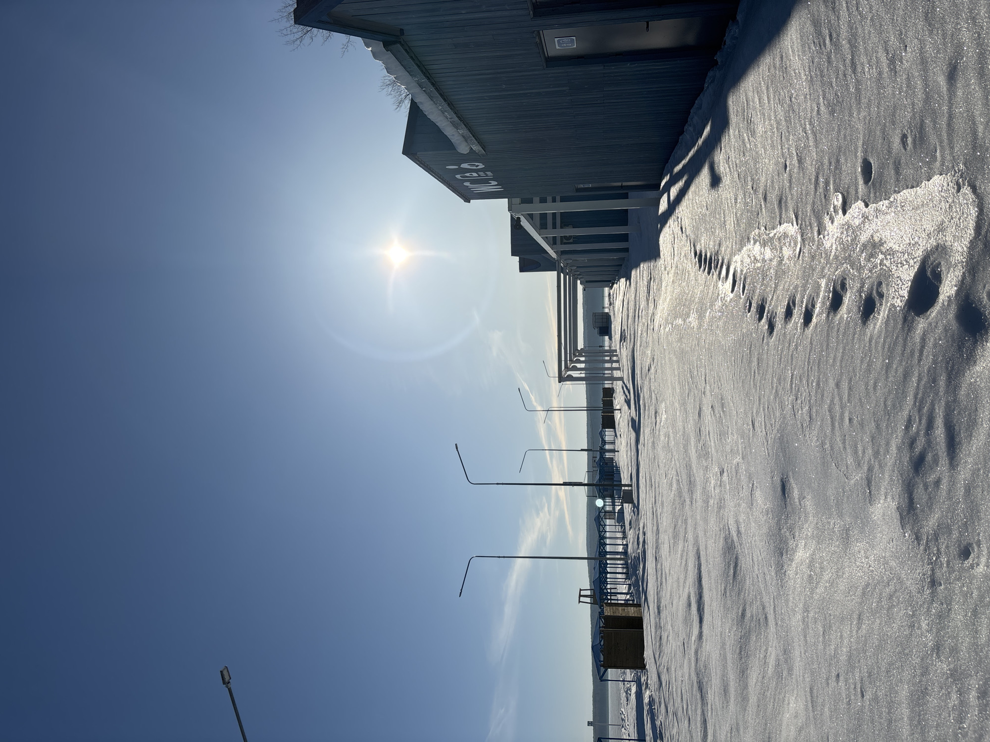 The image depicts a snow-covered pier or promenade in Chaikovsky, Russia on a sunny day. The view is oriented vertically, with the sky occupying a significant portion of the left side of the image. The sun is visible in the upper left, creating a bright glare. A long, low building lines the edge of the pier on the top side of the image. The building's facade is gray, and the name "NICE" is visible on it. Streetlights line the pier, extending into the distance. The ground is covered in snow, with footprints visible. In the distance, there is a body of water partially obscured by a slight haze. There are no people visible.