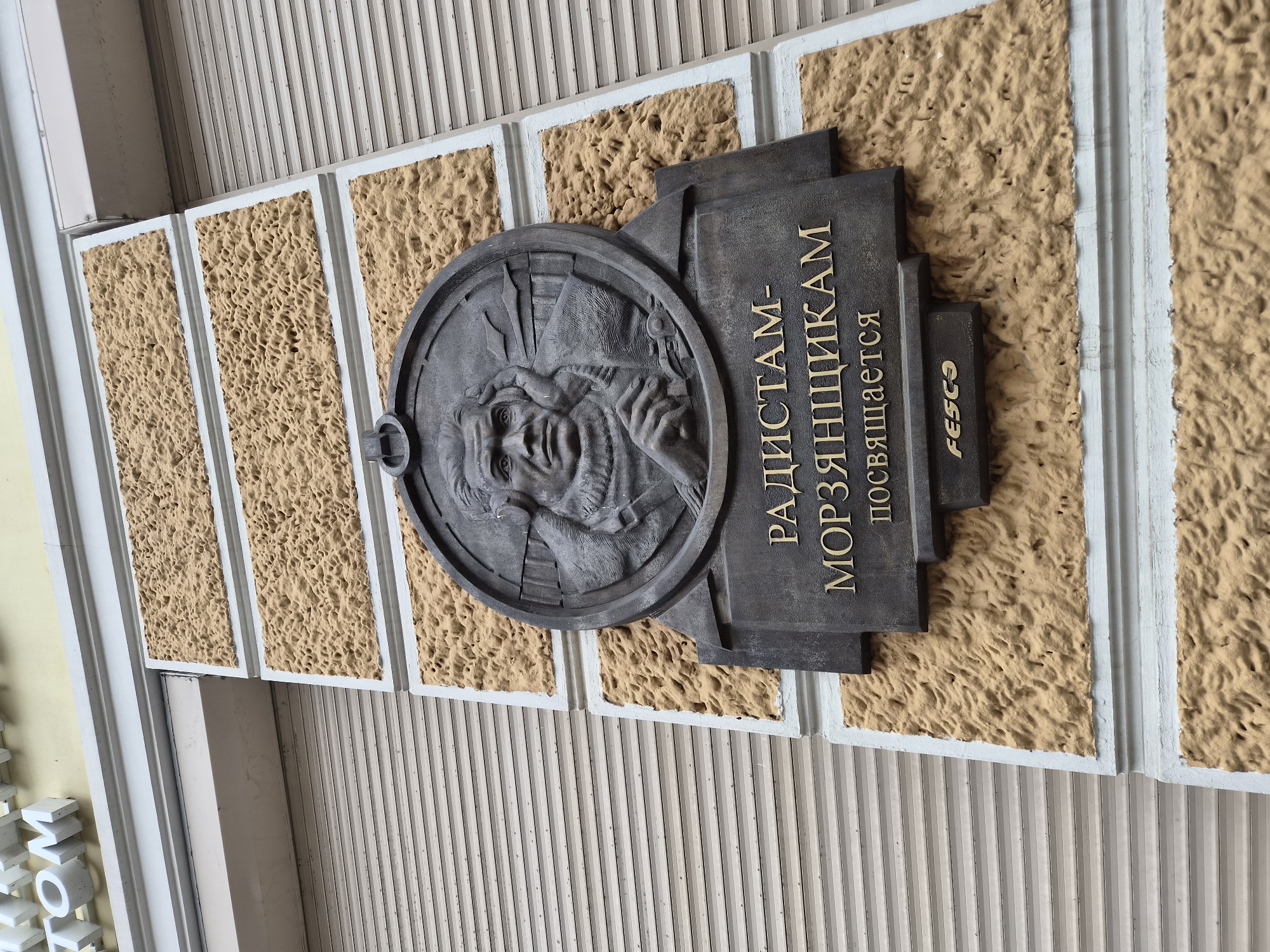 The image shows a relief plaque on the exterior wall of a building. The plaque is made of dark metal and features a portrait of a man in a circular frame. Below the portrait, there is an inscription in Cyrillic text and a logo that reads "FESCO". The text on the plaque reads: "РАДИСТАМ-МОРЗЯНЩИКАМ ПОСВЯЩАЕТСЯ" which translates to "Dedicated to radio operators-morzlyakshchikam". The building's facade is textured with a rough, sandy-colored material, bordered by white trim. A metal rolling shutter is visible on the right side of the image. The building appears to be located in Vladivostok, Russia, given the presence of the FESCO logo, which is a large transportation company headquartered in Vladivostok. The plaque is a memorial or dedication to radio operators.
