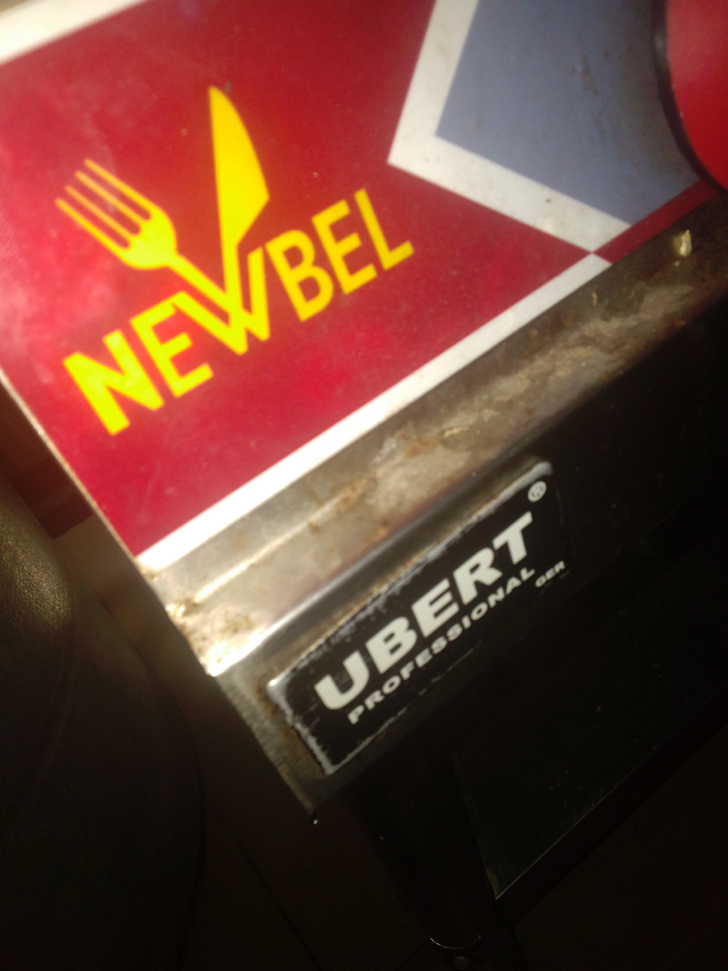 The image shows a close-up of a piece of kitchen equipment. The most prominent feature is a red panel with yellow lettering that reads "NEWBEL", accompanied by a yellow fork and knife graphic. To the right of this panel, there is a blue and white geometric design. Below the red panel, a metal plate with text is visible. The text on this plate reads "UBERT PROFESSIONAL GER". The overall scene appears to be indoors, likely in a kitchen or food service environment, given the branding. The equipment looks somewhat used, with signs of wear and possibly some rust on the metal trim. The lighting is dim, suggesting it might be evening or that the area is not brightly lit. There are no people visible in the image. The context of Howayi, Nigeria, provides a potential geographical location for this equipment.