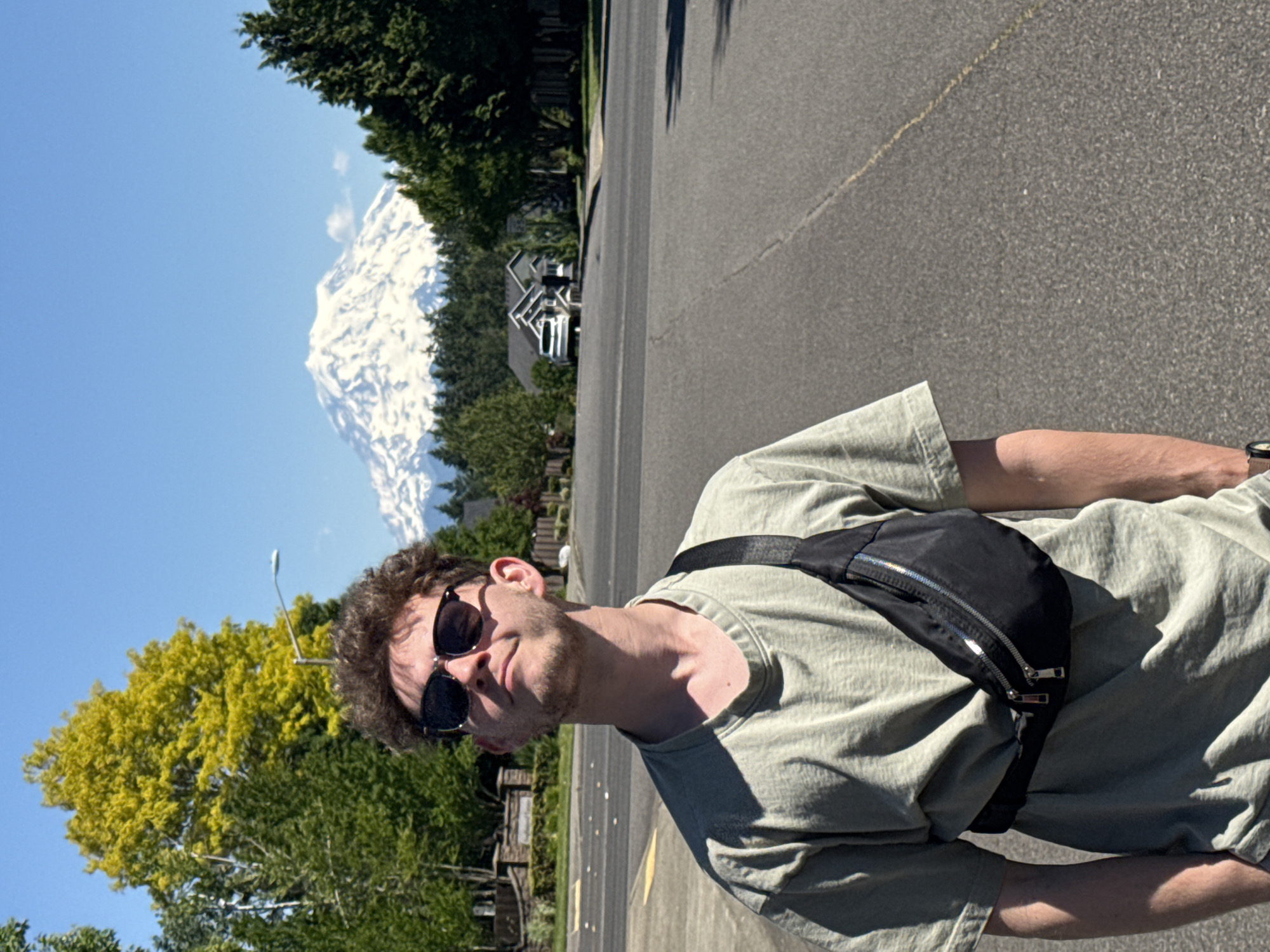 A young man with curly brown hair and sunglasses stands in the middle of a paved road in South Hill, USA, with a snow-capped mountain in the background. He is wearing a light green t-shirt and a black fanny pack across his chest. The sun is shining brightly, casting shadows on the road and the man. Trees and houses are visible on either side of the road. The mountain is large and imposing, with snow covering its peak and slopes. The sky is a clear blue.