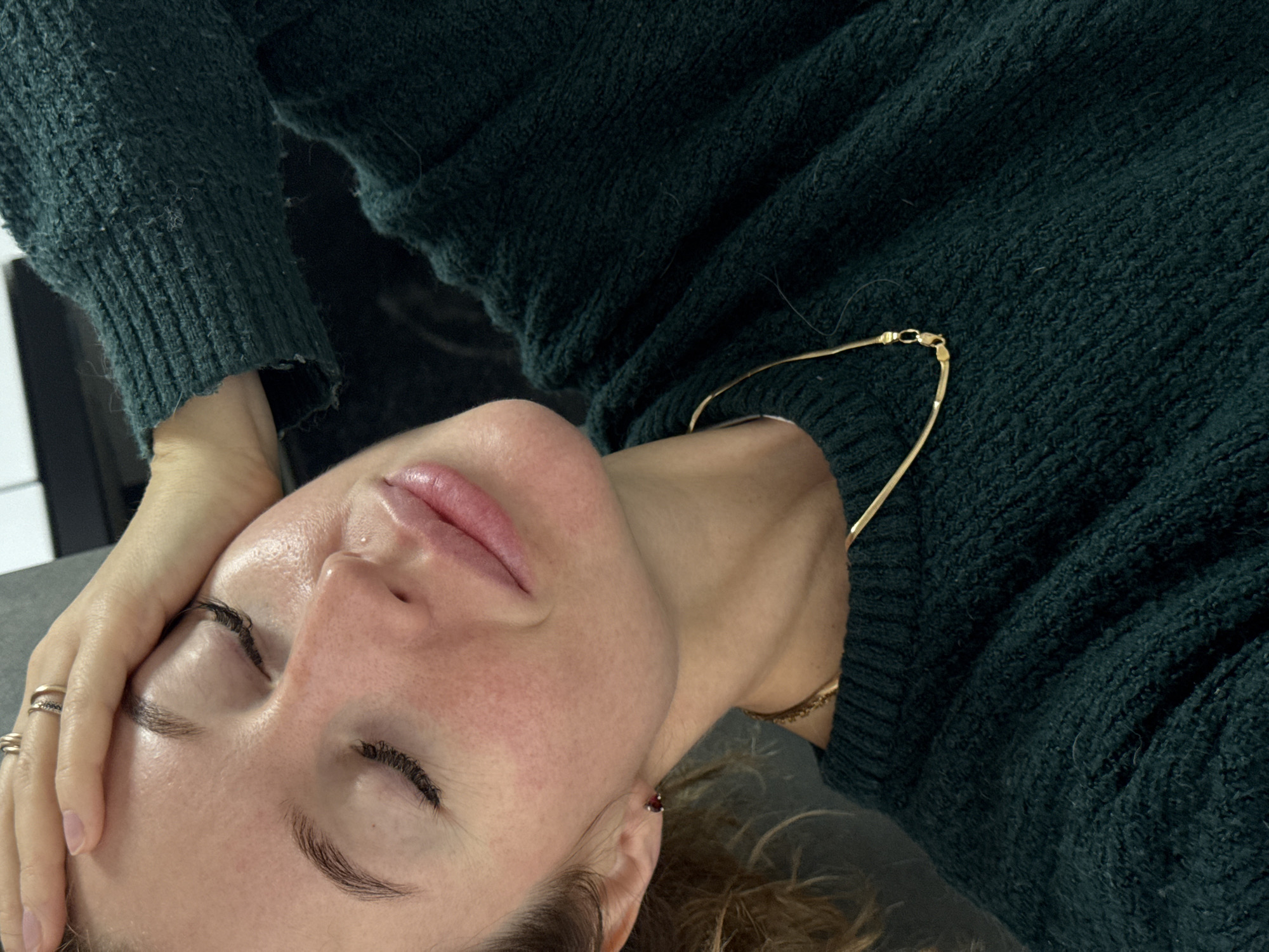 A fair-skinned woman with closed eyes, a pierced nostril, and several rings on her left hand is seen in a close-up, tilted perspective. She is wearing a dark green, textured sweater and a delicate gold chain necklace. The background consists of a grey surface and a hint of a white object, possibly a piece of furniture. There is no evidence to determine if the image was taken in the city of Chaikovsky, Russia.