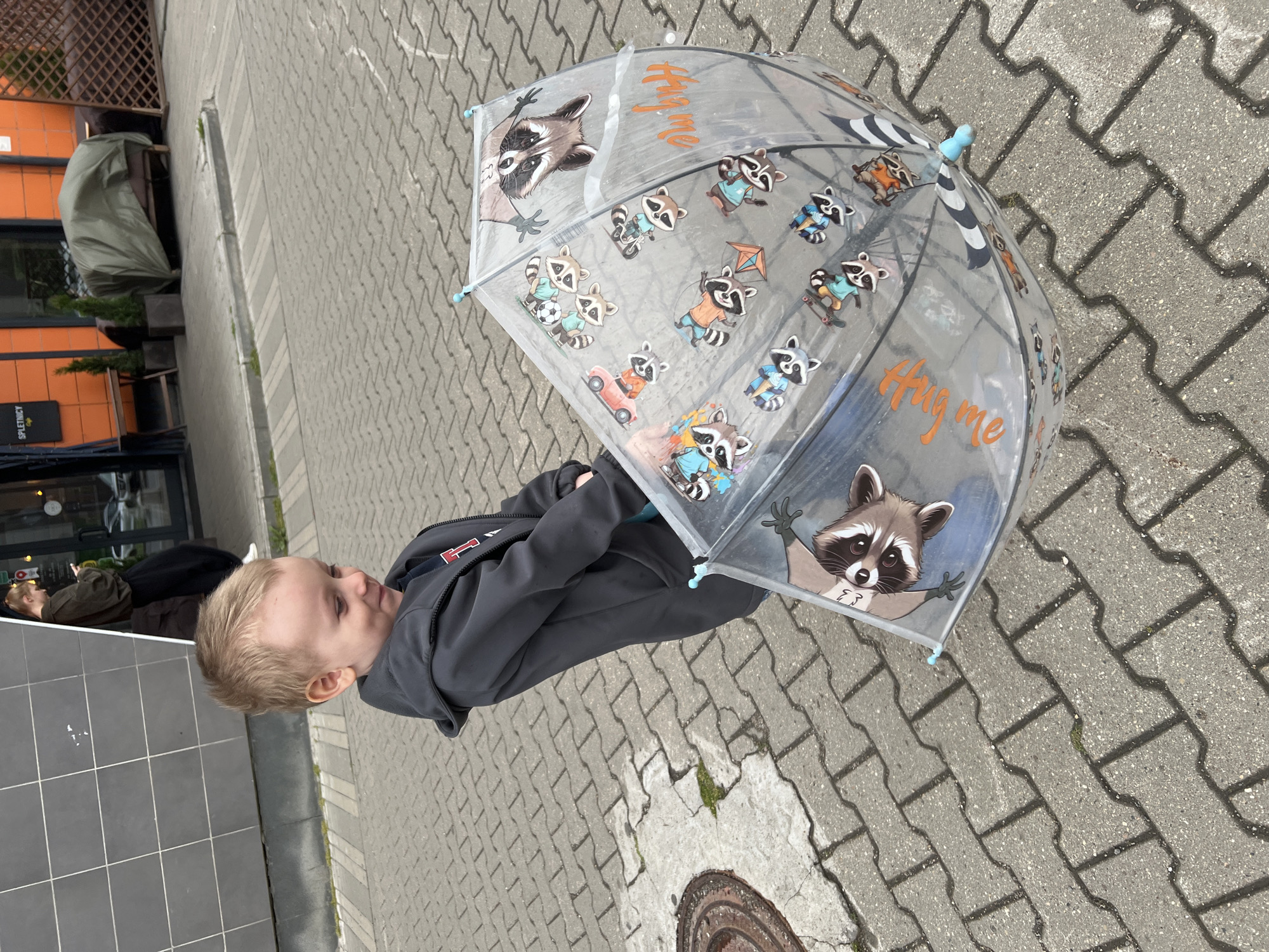 A small blond boy in a gray hooded sweatshirt stands on a gray brick sidewalk, holding a transparent umbrella decorated with raccoon illustrations. The umbrella features the words "Hug me" in orange lettering. The boy is looking to the side. Behind him is a building with an orange facade and a black entrance area. A green object covered with a tarpaulin is visible near the building. In the foreground, there's a manhole cover embedded in the sidewalk. The location is likely a street in Mytishchi, Russia, based on the provided information.