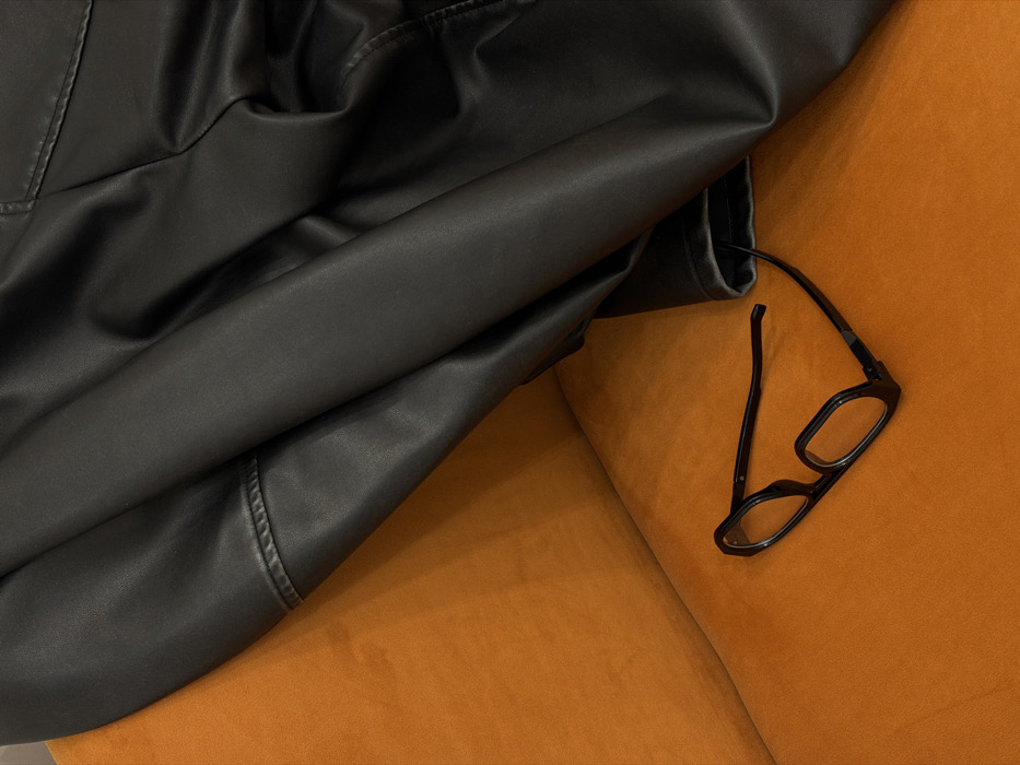 A dark, folded garment, resembling a jacket, lies across the upper right and central areas of an orange upholstered surface. A pair of black-framed eyeglasses with rectangular lenses, one temple extended, rests on the lower-left portion of the same surface. The indoor scene is evenly lit. This depiction is set in Yerevan, Armenia.