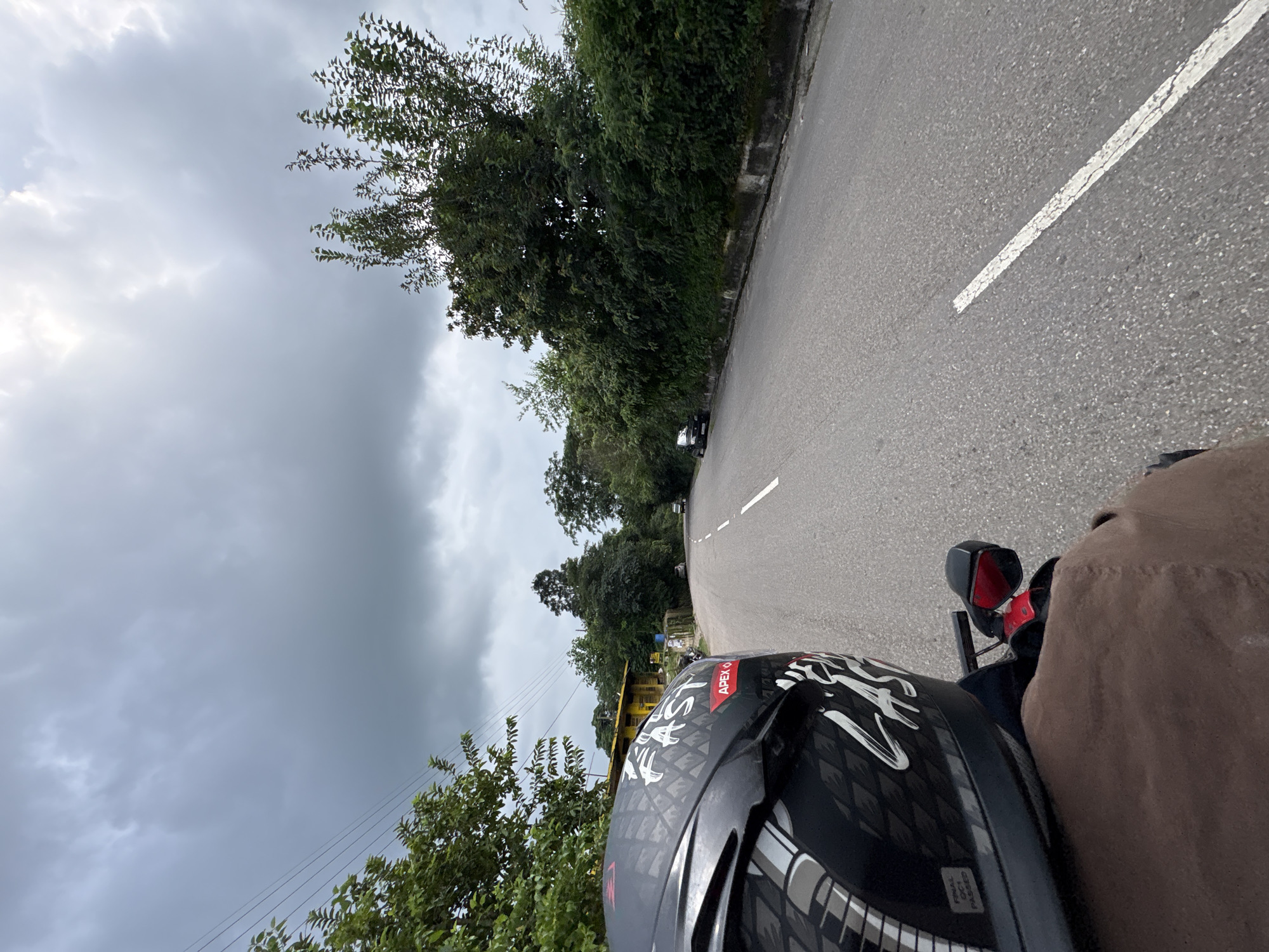 The image depicts a point-of-view shot from a motorcycle traveling on a paved road. The road is lined with lush green trees and vegetation on both sides. In the distance, there are a few buildings, including a yellow structure with a red roof. The sky is overcast with dark, heavy clouds, suggesting impending rain. A car is visible further down the road. The foreground shows the rider's helmet, which is black with a patterned design and text that includes "FAST" and "APEX 6". The rider's brown jacket is also visible. The location is identified as Baldwara, India.