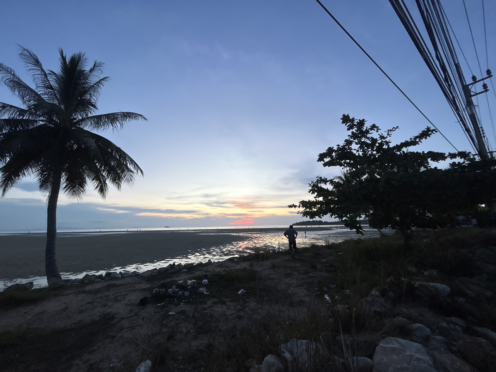 The image depicts a coastal scene at dusk, likely in Baan Sri Thanu, Thailand. A silhouetted palm tree stands on the left, contrasting with a tree and overhead power lines on the right. A person is visible standing near the shoreline, appearing to look out towards the water. The beach is wide and flat, with pools of water reflecting the fading light of the sunset. The foreground is darker, showing rocks and vegetation. The sky is a gradient of blues and pinks, indicating the end of the day.