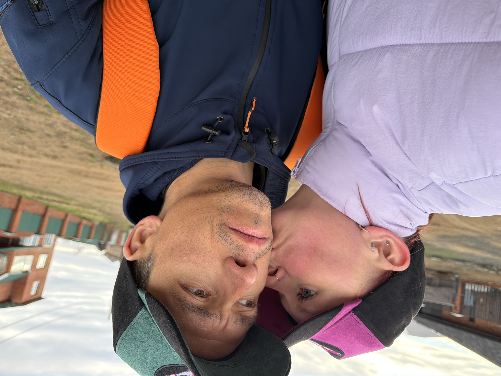 This vertically oriented image captures two individuals, a man and a woman, in an intimate and affectionate close-up. The man, positioned on the left side of the frame, has light skin, noticeable stubble, and brown eyes, looking towards the viewer with a thoughtful expression. He is wearing a dark blue jacket with bright orange accents on the shoulders and a black zipper with an orange pull. His head is adorned with a baseball cap featuring dark green/teal and black panels.
The woman, on the right, has light skin and light-colored eyes, gazing subtly away from the camera. She wears a light purple puffy jacket and a baseball cap with black and magenta/pink sections. Her ear is visible, showcasing multiple piercings, and she has a silver nose ring. Her face is pressed gently against the man's cheek or temple, implying a tender kiss or nuzzle.
In the background, visible above their heads, is a residential or urban scene featuring buildings with red-brown brick walls and green roofs, under a partly cloudy sky. This architectural style provides a location cue consistent with the specified context of Chaikovsky, Russia. The lighting suggests it is daytime, possibly overcast or late afternoon. The overall emotion conveyed is one of warmth and affection between the subjects. There is no visible text in the image.