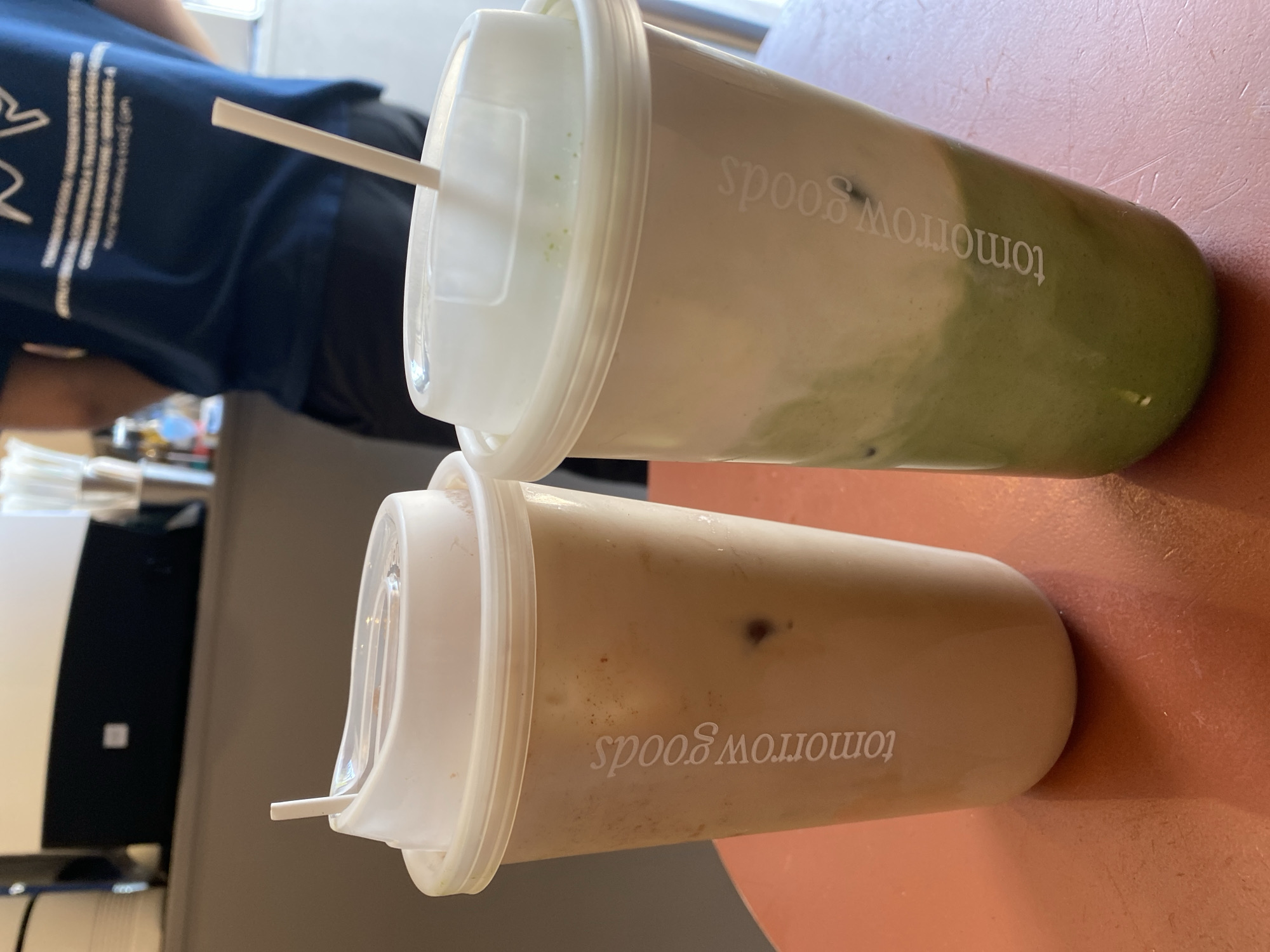 The image displays two cold beverages in plastic cups with white lids and straws, positioned on a reddish-brown table. The photograph is oriented sideways, rotated 90 degrees counter-clockwise. A person, partially visible in the blurred background, is wearing a dark blue t-shirt with an indistinct white graphic design or text on the back.
The upper cup contains a visually appealing layered drink, with a bright green base (suggesting matcha or a similar flavored beverage) topped with a creamy white layer. A white plastic straw protrudes from its translucent white lid. The side of this cup clearly displays the white text "tomorrow goods", appearing sideways and inverted relative to the image's rotation. Some small green speckles are visible inside the lid.
Below it, the second cup holds a uniform light brown or beige beverage, likely coffee or milk tea, with a few ice cubes discernible within. This cup also has a white plastic straw and a translucent white lid, and the same "tomorrow goods" text is printed on its side.
The table surface has a warm, reddish-brown hue. The blurred background indicates an indoor setting, possibly a cafe or restaurant, where these drinks have been served. The overall scene suggests a casual moment, perhaps during a break or meal, in Puebla, Mexico, as provided by the context. The lighting appears to be natural daytime light.