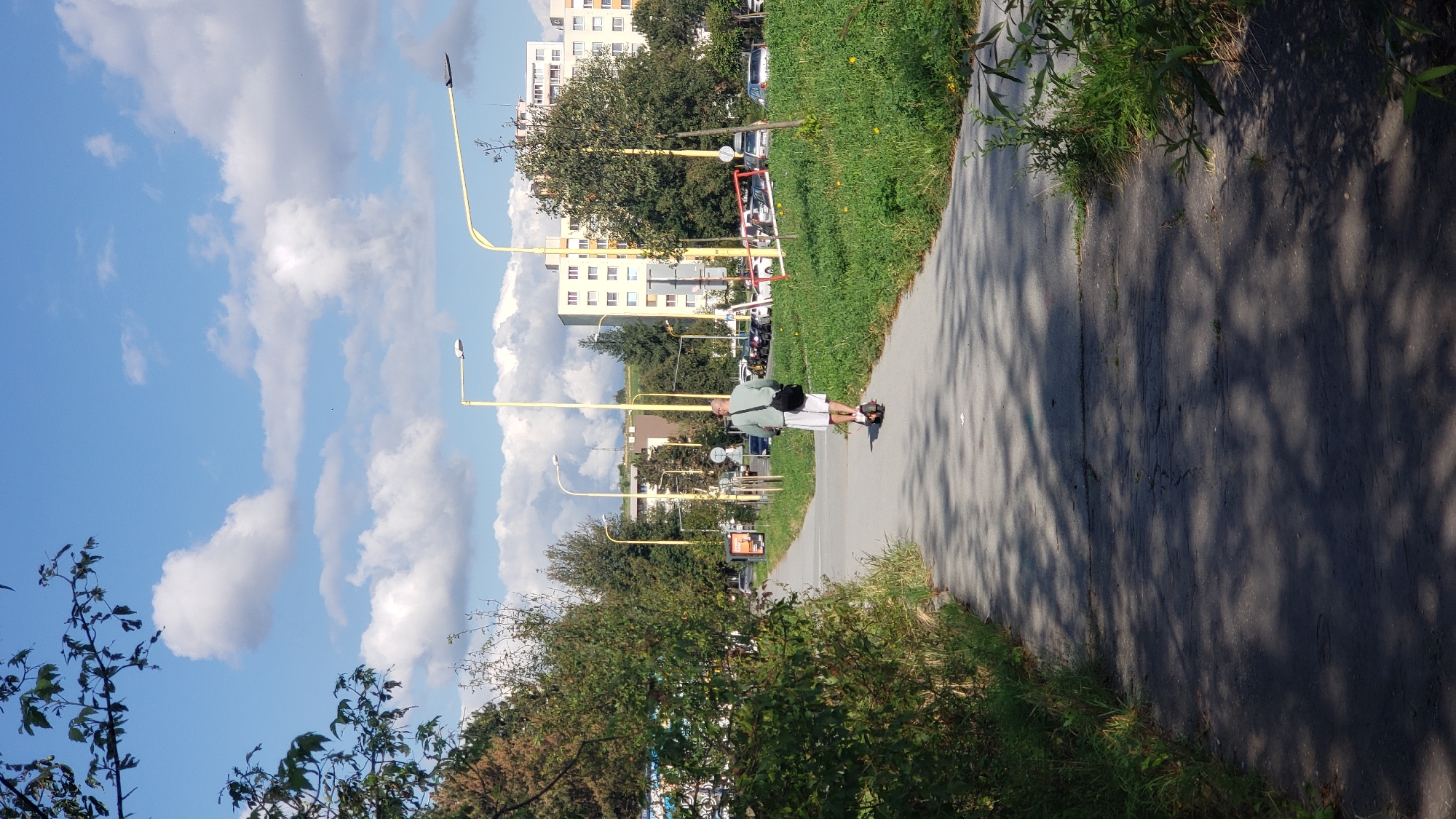 A person is riding an electric scooter down a paved pathway in Prešov, Slovakia. The person is bald, wearing a light green t-shirt and white shorts, and carrying a black bag over their shoulder. The pathway is lined with green grass and trees on either side. In the background, there are several apartment buildings and cars parked in a lot. Yellow streetlights are visible along the pathway. The sky is blue with scattered white clouds.
