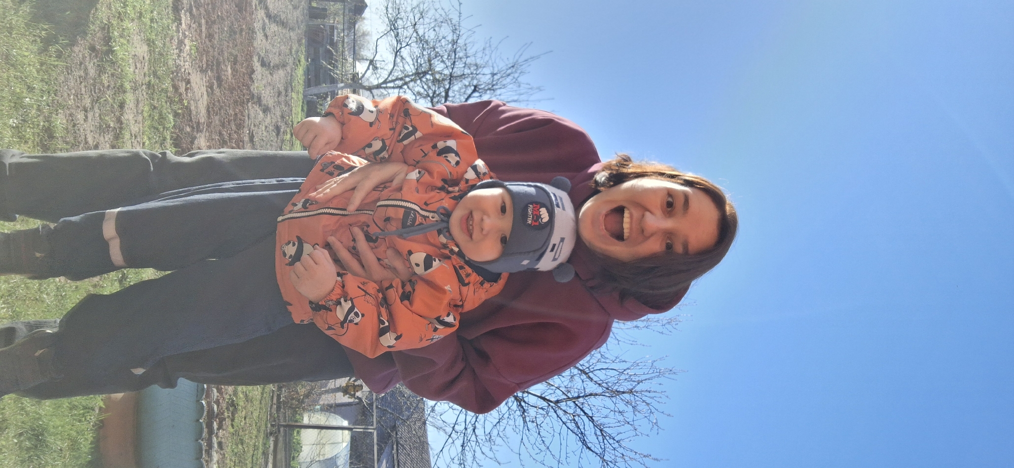 The image captures a woman holding a young child in an outdoor setting. The woman is wearing a maroon-colored hoodie and dark pants. She is smiling widely with her mouth open. The child is bundled in an orange jacket patterned with panda designs and a blue hat with a graphic print. They both appear to be looking directly at the camera. The background includes a clear blue sky, a bare tree, grass, and the edge of a building or structure. The location is possibly in Chaikovsky, Russia.