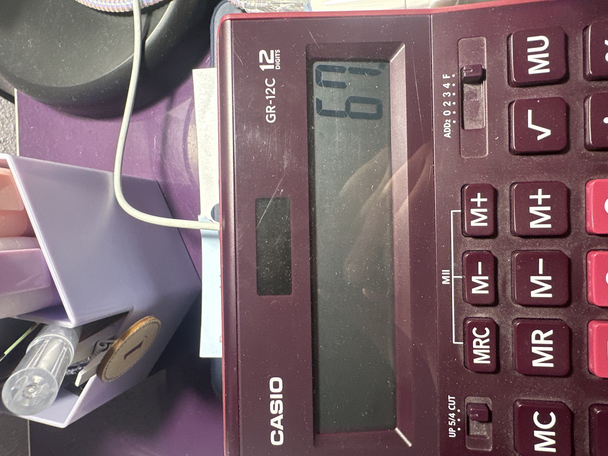 A burgundy Casio GR-12C calculator with a 12-digit display is prominently featured in a close-up, top-down shot. The calculator's screen shows the number "67". The device's surface appears slightly dusty and has minor scratches. The brand "CASIO" is visible near the bottom left, and "GR-12C 12 DIGITS" is printed above the display.
Several buttons and switches are visible on the calculator. The keypad includes buttons labeled "MU", "√", "M+", "M-", "MRC", "MR", and "MC". There are two slider switches: one labeled "ADD2 0 2 3 4 F", which is currently set to "2", and another labeled "UP 5/4 CUT".
The calculator rests on a solid purple surface. To its left, a light purple desk organizer contains a clear cylindrical object and a round, light-colored object with a central marking, possibly a wooden token. A white cable runs across the upper left part of the image. The scene suggests an office or workspace setting.