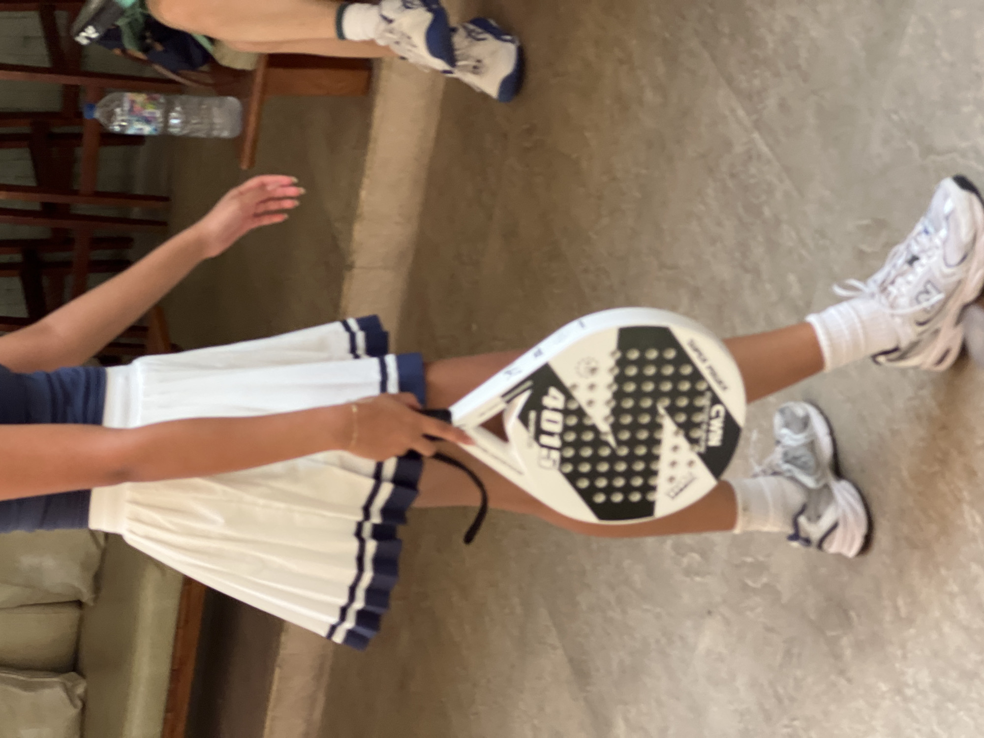 A person wearing a navy blue top, a white pleated skirt with navy trim, white socks, and white athletic shoes is holding a white paddle tennis racket. They are standing on a light-colored tiled floor. In the background, there is a light-colored sofa with pillows, wooden chairs, and a bottle of water on a wooden bench. The scene appears to be indoors, possibly in a recreational facility or lounge area, in the city of North Kuta, Indonesia.