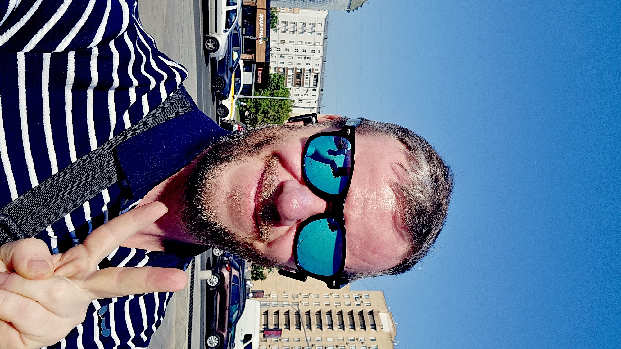 A selfie shows a person wearing sunglasses and a blue-and-white striped shirt making a peace sign with their left hand. The sunglasses reflect a building and sky. The background includes other buildings and vehicles, and a clear blue sky. Based on architectural style and other elements, the location is likely Moscow, Russia.