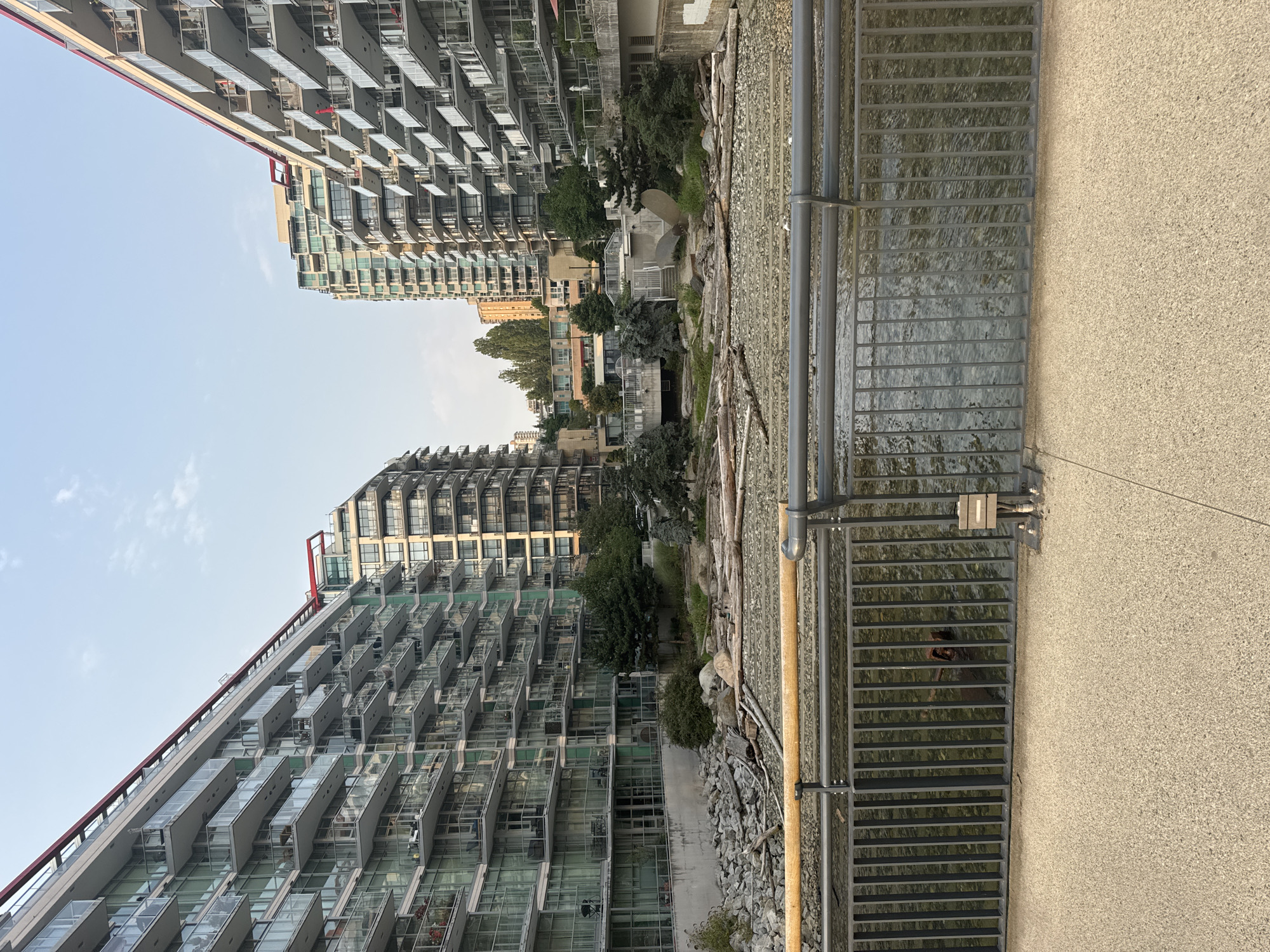 The image depicts an urban waterfront scene in North Vancouver, Canada, likely within the area of the False Creek or Burrard Inlet waterfront developments, characterized by modern high-rise residential buildings. Two prominent apartment complexes, one on the left and one on the right, feature extensive glass balconies with metal railings, suggesting a contemporary architectural style. Between these buildings, a dry riverbed or foreshore is visible, filled with rocks and driftwood. A metal railing runs horizontally across the foreground, separating a concrete walkway from the water below. The water is shallow and appears to be a mix of natural flow and possibly tidal influence. The sky is partly cloudy.