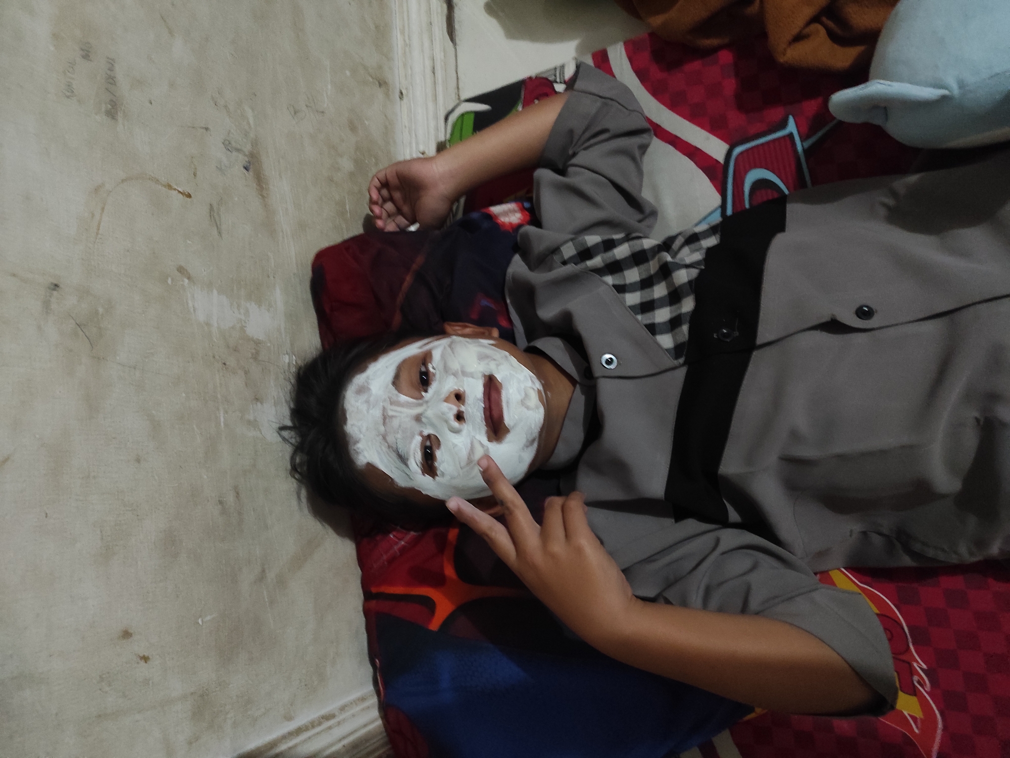 A young person lies on their back with a white face mask applied to their face. They are wearing a grey and black checkered shirt over a grey shirt. They are lying on a bed with a colorful patterned comforter. The background appears to be a wall with some writing on it. The image was taken in the city of Sumedang, Indonesia.