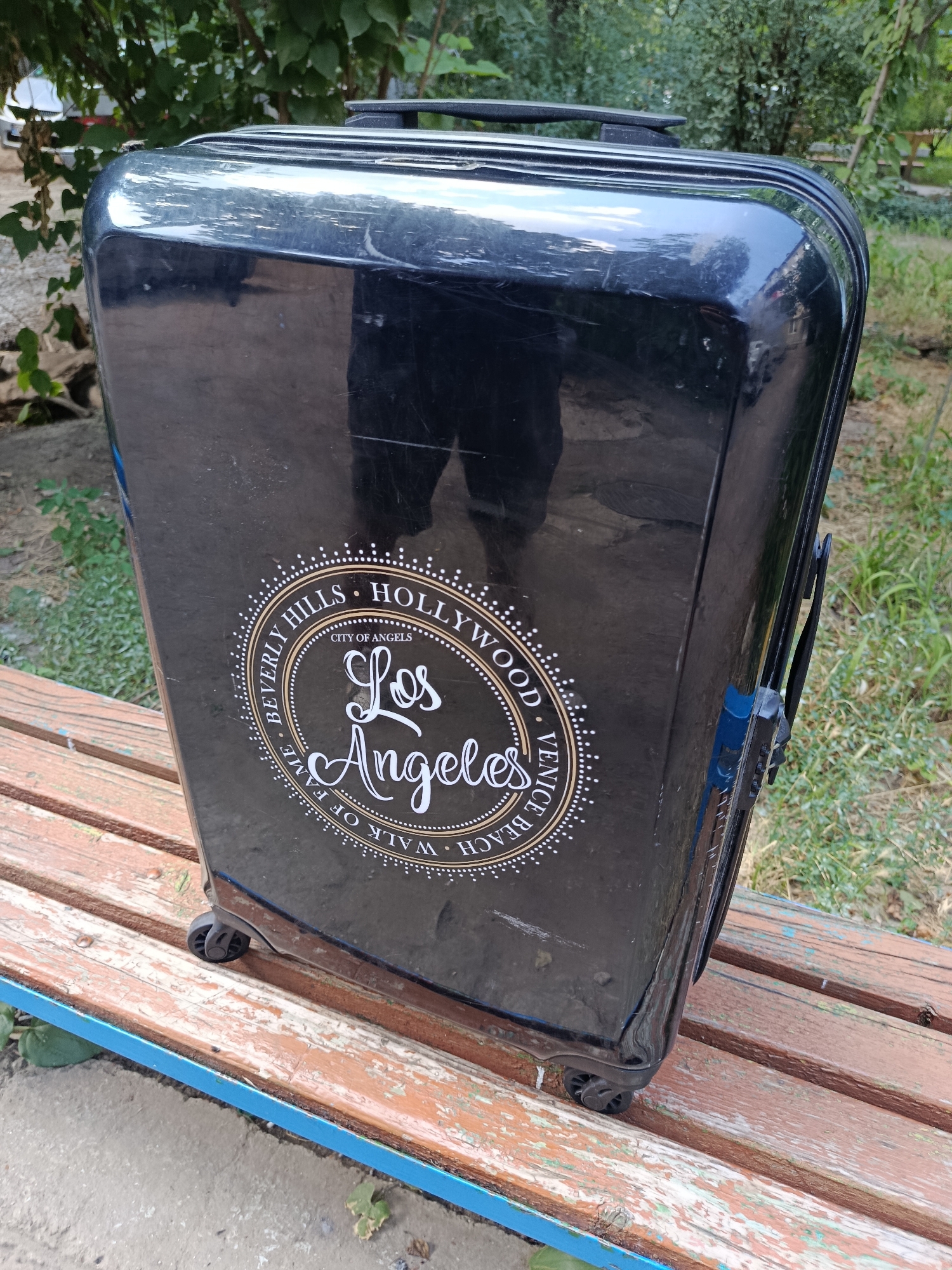A black suitcase with a circular sticker featuring "Los Angeles" and other California landmarks rests on a weathered wooden park bench with blue trim. The suitcase has visible scuff marks and is equipped with wheels. The background consists of green foliage and trees, suggesting an outdoor setting, possibly a park. No people are present in the image. The scene is captured in daylight.