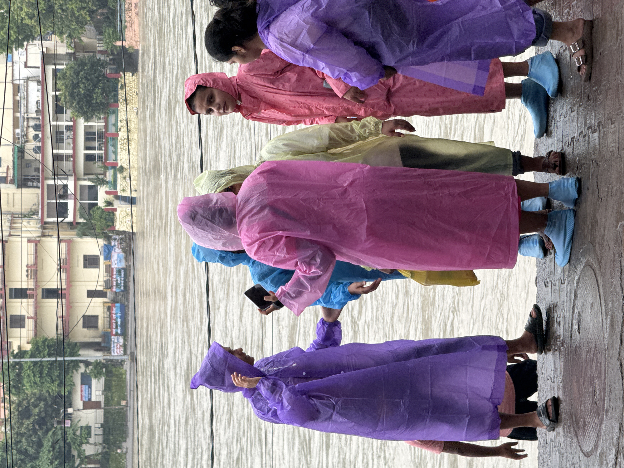 Six individuals, some wearing colorful raincoats and boots, stand on a wet paved surface near a wide, muddy river. In the background, buildings with multiple stories and balconies are visible, with trees and power lines interspersed. The river appears to be at a high level, possibly due to rain, and the sky is overcast, suggesting a rainy day in Rishikesh, India, likely near the banks of the Ganges River. One person in a purple raincoat is holding up a smartphone, possibly taking a picture or video. Another person in a pink raincoat is looking towards the camera. The ground is wet, with puddles reflecting the surroundings.