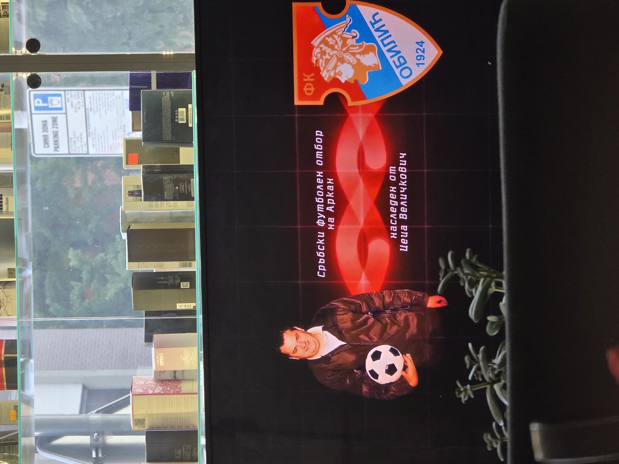 The image depicts a retail display, likely in a shopping mall or department store in Varna, Bulgaria. The upper portion shows a shelf filled with various perfume or cosmetic boxes, some branded "Versace". Behind the shelves, a parking zone sign is visible. The lower portion displays a large screen showing a man holding a soccer ball. Text in Cyrillic script, likely Serbian or Bulgarian, appears on the screen above the man, and a shield-shaped logo with Cyrillic text and the year "1924" is to the right. In the foreground, small potted plants are visible on a dark surface, possibly a counter or display. The scene appears to be indoors, illuminated by artificial lighting.
