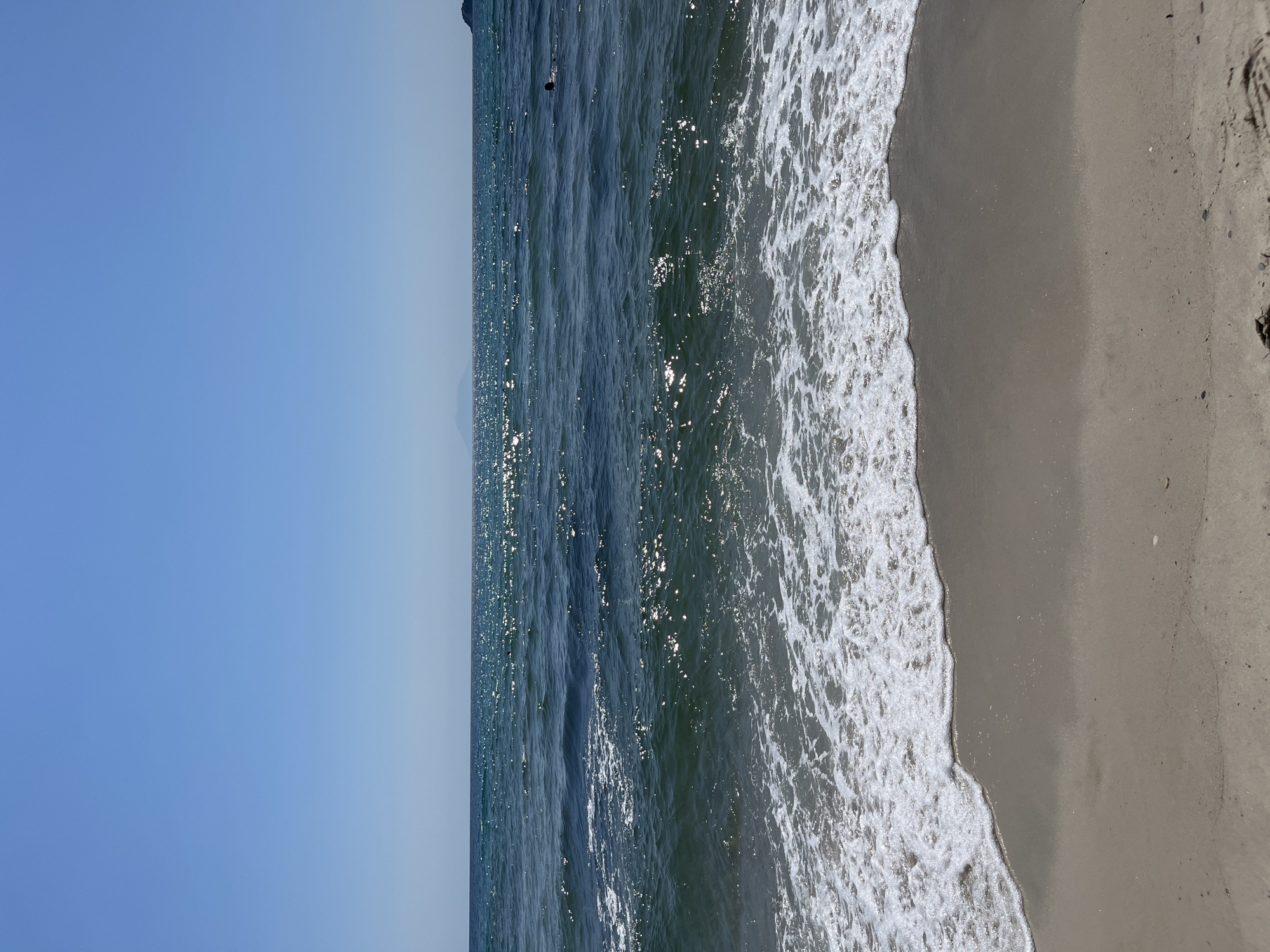 The image shows a clear blue sky over a calm ocean with sparkling sunlight reflecting on the water. Gentle waves are lapping at a sandy beach in the foreground. In the distance, a faint outline of mountains can be seen on the horizon. A single person is swimming in the ocean on the right side of the frame. The overall scene is serene and idyllic, evoking a sense of peace and relaxation. The location is Unknown, Unknown.