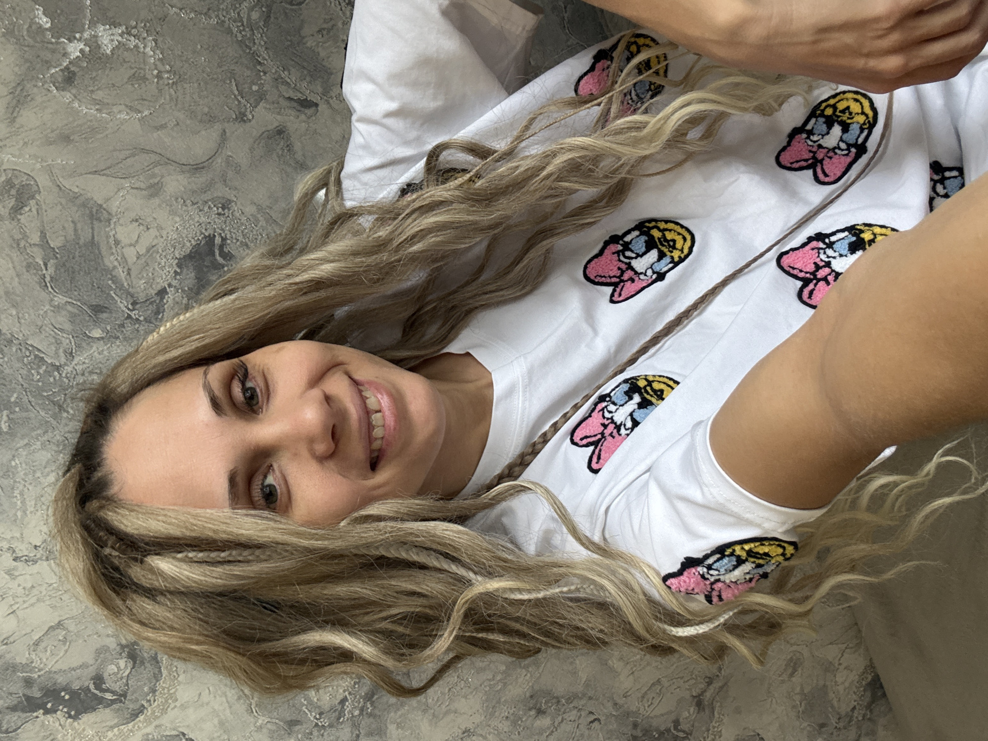 The image shows a woman lying on what appears to be a sofa or bed. She has long, wavy, blonde hair with some braids visible. She is smiling and looking directly at the camera. She is wearing a white t-shirt with cartoon-like patches scattered across it. The background includes a textured grey surface that could be a wall or another part of the furniture. The location cannot be determined from the information provided.