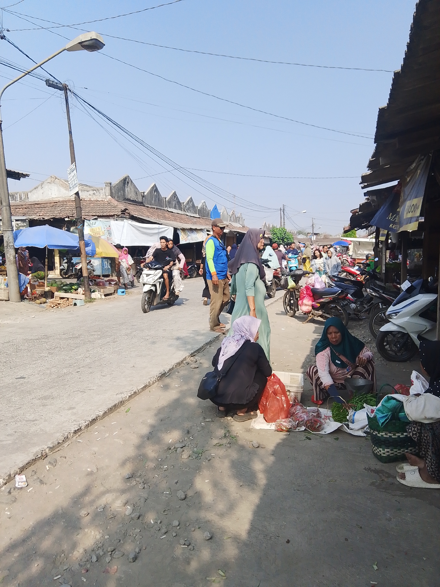 The image depicts an outdoor marketplace scene in the city of Kalinyamatan, Indonesia. The foreground shows several women squatting on the ground, engaged in selling produce. One woman, wearing a dark green headscarf, is arranging green vegetables. Another woman, in a patterned headscarf, is sitting behind a pile of produce, and a third woman is partially visible in the lower left, also with a headscarf. Various bags and baskets filled with produce are scattered around them.
In the middle ground, a white motorcycle is being ridden by a man and a woman, both wearing casual clothing and headscarves. Behind them, other market stalls and people are visible. A blue umbrella and a yellow umbrella are set up, indicating vendor stalls. Numerous motorcycles are parked or in use, suggesting the common mode of transportation.
The background reveals a street lined with buildings that appear to be shops or market structures, with angled rooftops. Power lines crisscross the sky, and a lamppost is visible on the left side of the image. The overall environment suggests a busy, sunny day at a local market. The ground is paved with concrete in some areas and dirt with scattered debris in others.