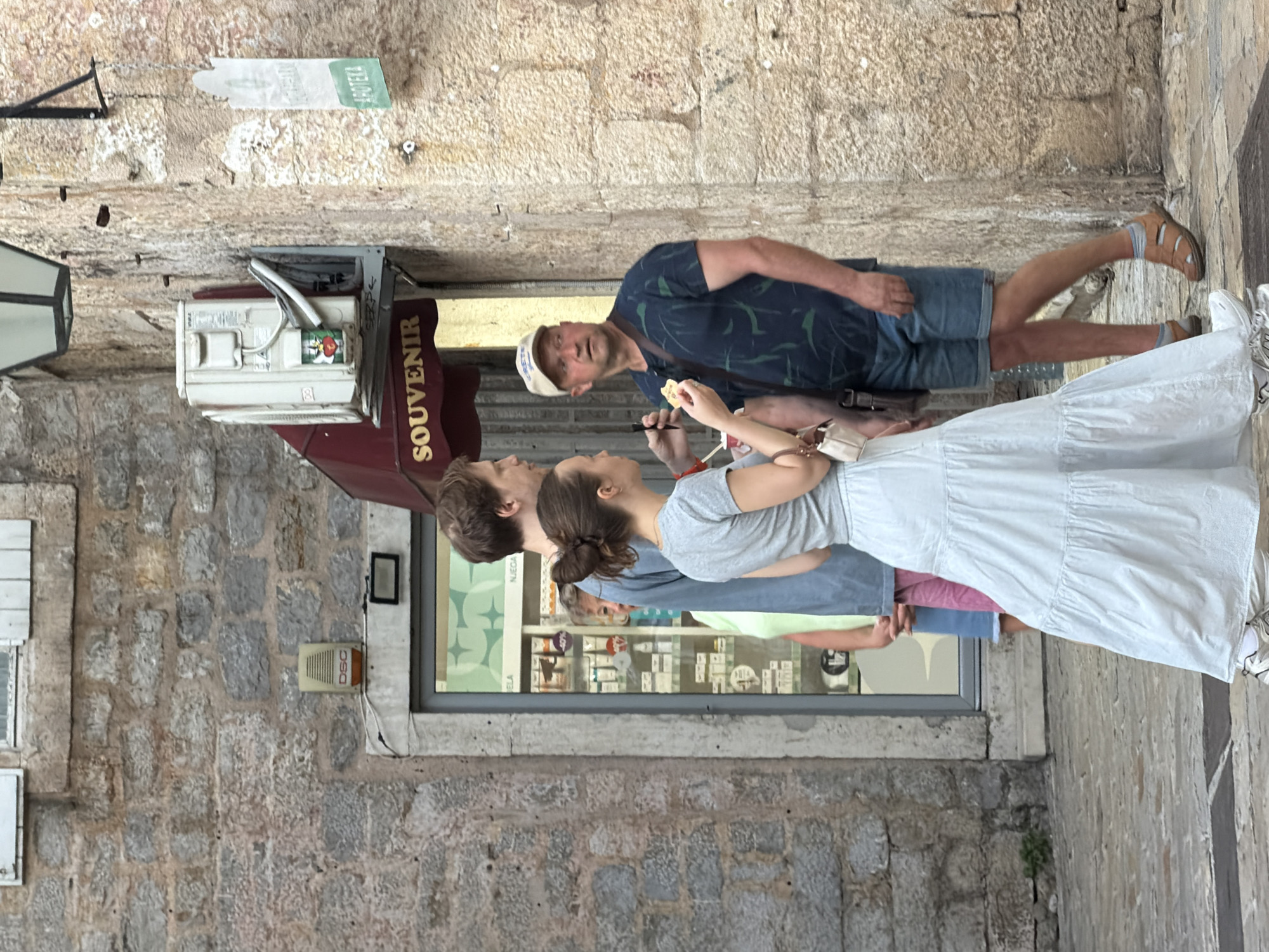 In Budva, Montenegro, four individuals are present on a cobblestone street in front of a building with a stone facade. The building features a shop with a glass entrance displaying merchandise. Above the shop entrance is a burgundy awning with the word "SOUVENIR" in gold lettering. To the right of the awning, an air conditioning unit is mounted on the wall. Further to the right, a sign for "APOTEKA" (pharmacy) is visible.
In the foreground, a woman wearing a light blue tiered maxi skirt and a grey t-shirt stands facing left, holding a small white purse. She is partially obscuring a man to her left, who is wearing a blue shirt and also facing left. Behind these two, a person in a light green top and pink shorts is visible in the shop entrance. To the right of the woman in the blue skirt, a man wearing a dark blue t-shirt with a leaf pattern, blue denim shorts, a beige baseball cap, and brown sandals is looking to his right. He is holding a small piece of food. A dark brown strap is visible across his chest. The overall scene suggests a shopping area in Budva's old town.