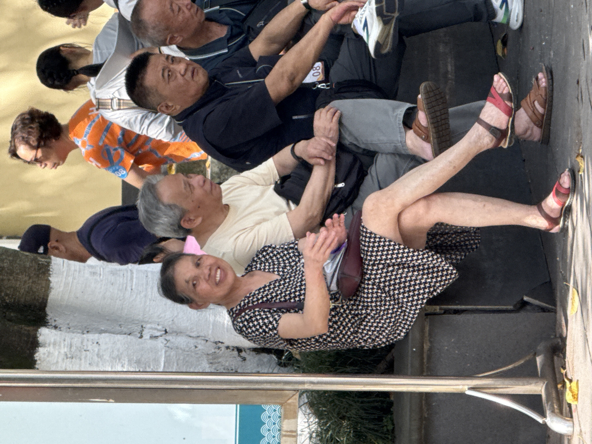 A group of people, presumably tourists, are situated in an outdoor location in Jiulongpo District, China. Individuals are seated and interacting. One older woman in the foreground is smiling and wearing a patterned dress with sandals. Several other people, including some wearing running gear, are arranged behind her. The environment includes urban infrastructure like railings and signage.