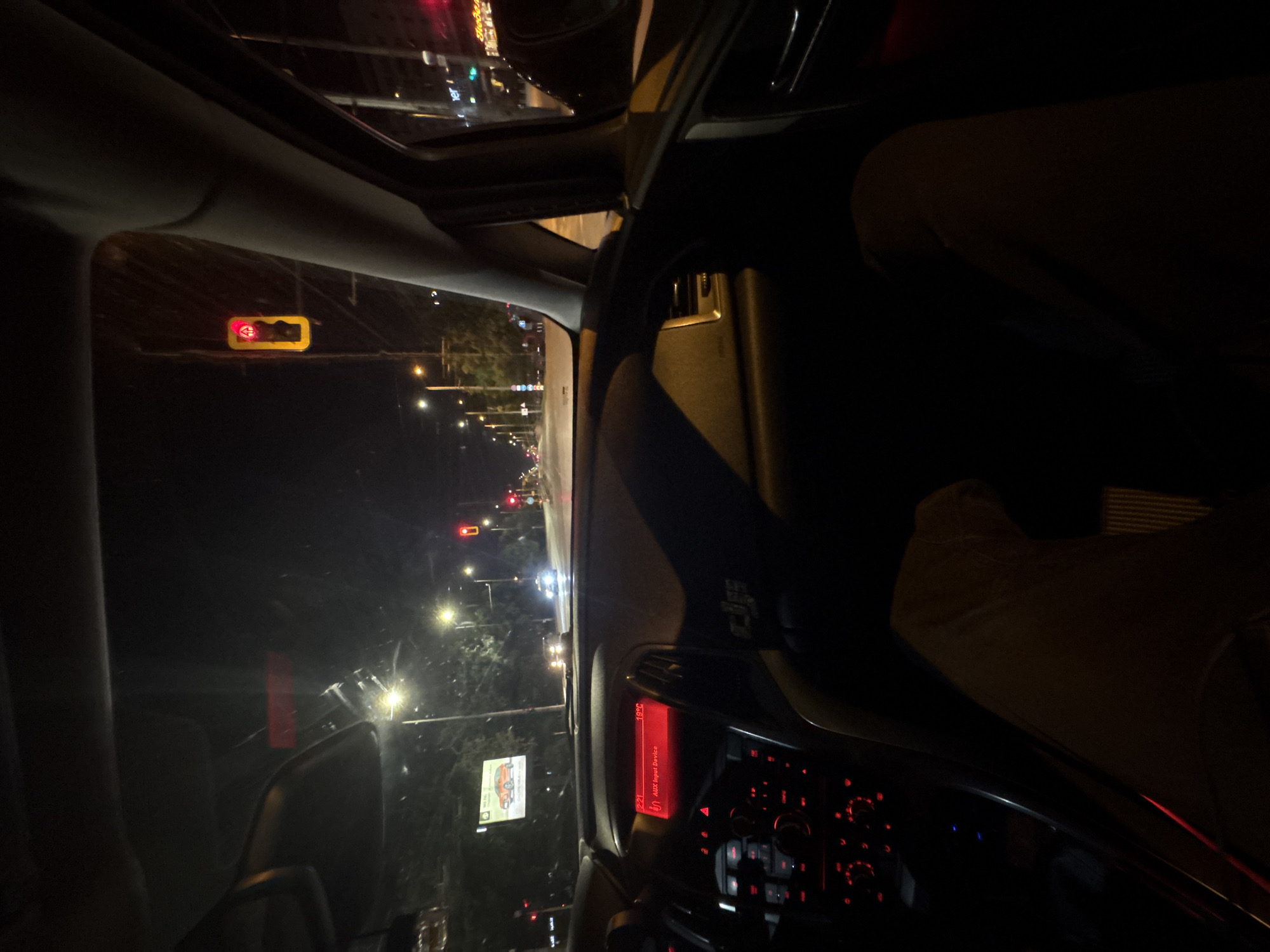 The image, taken from inside a car at night, shows a view of a city street in Sofia, Bulgaria. The car is stopped at a red traffic light. The dashboard of the car is visible, illuminated with red lights, and the display shows "19.5C" along with "AUX Input Device." Out the window, there are streetlights and other cars on the road. A billboard with an image of a red car is also visible. The scene is dark, typical of a nighttime urban environment.