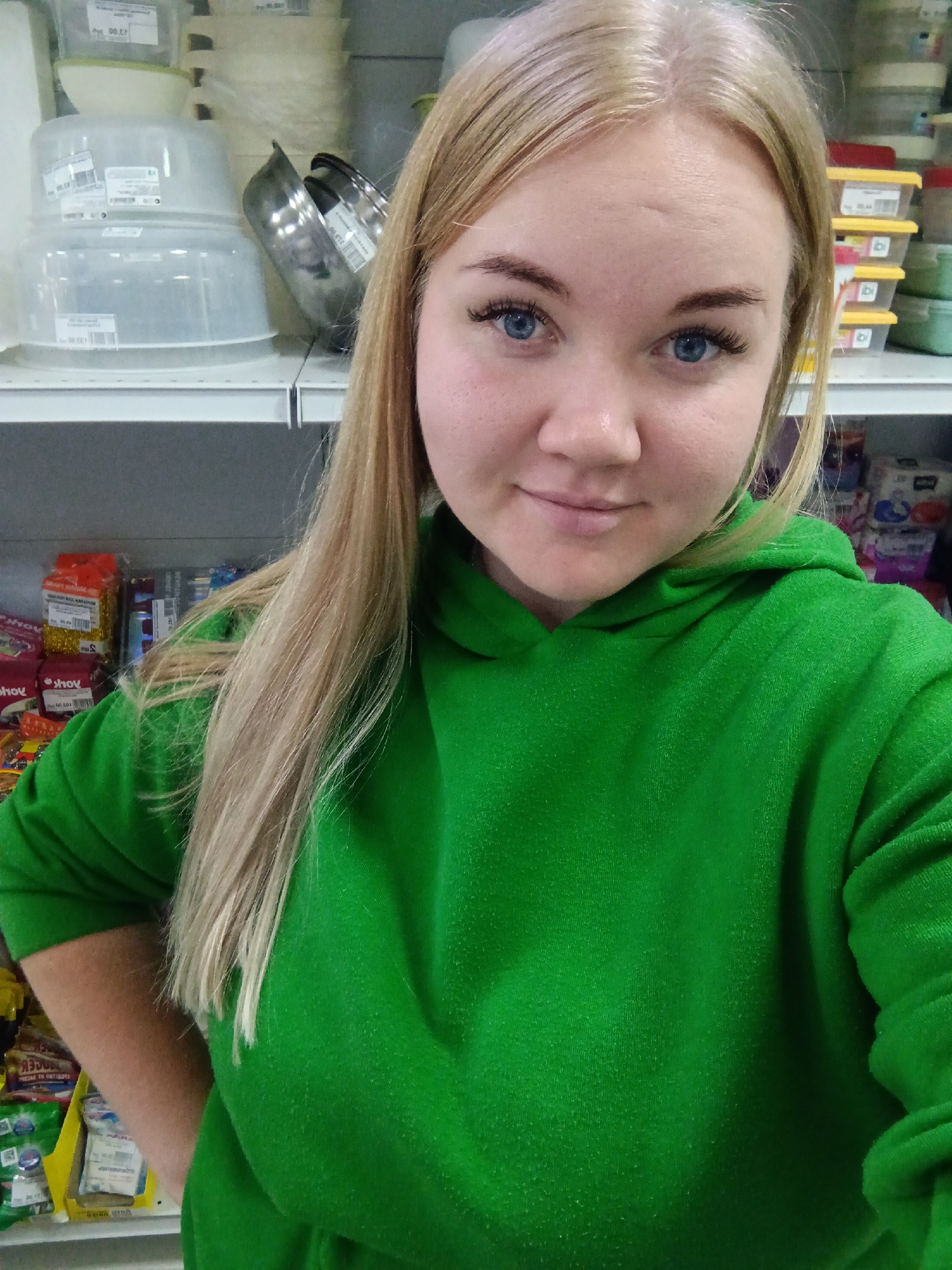 A young woman with long, straight blonde hair and striking blue eyes is featured in a close-up, chest-up portrait. She has fair skin and is wearing a vibrant green hooded sweatshirt (hoodie). Her expression is calm and friendly, with a slight, demure smile, and she is looking directly at the camera. Her eyelashes are notably long.
The setting appears to be an indoor retail or storage area, consistent with a shop or warehouse environment, located in Foki, Russia. In the background, shelves are stocked with various items. On the upper left, clear plastic containers, possibly for food or household storage, are stacked, some bearing labels with indistinct text and numbers like "00.ET" and "00.12". Next to these are a few metal bowls. To the right, shelves hold smaller containers with yellow lids and other packaged goods, some wrapped in red and yellow materials, and some displaying reversed Cyrillic text, such as "shov". The lighting is artificial, typical of an indoor commercial space. The woman is posing for the photograph, likely a selfie.