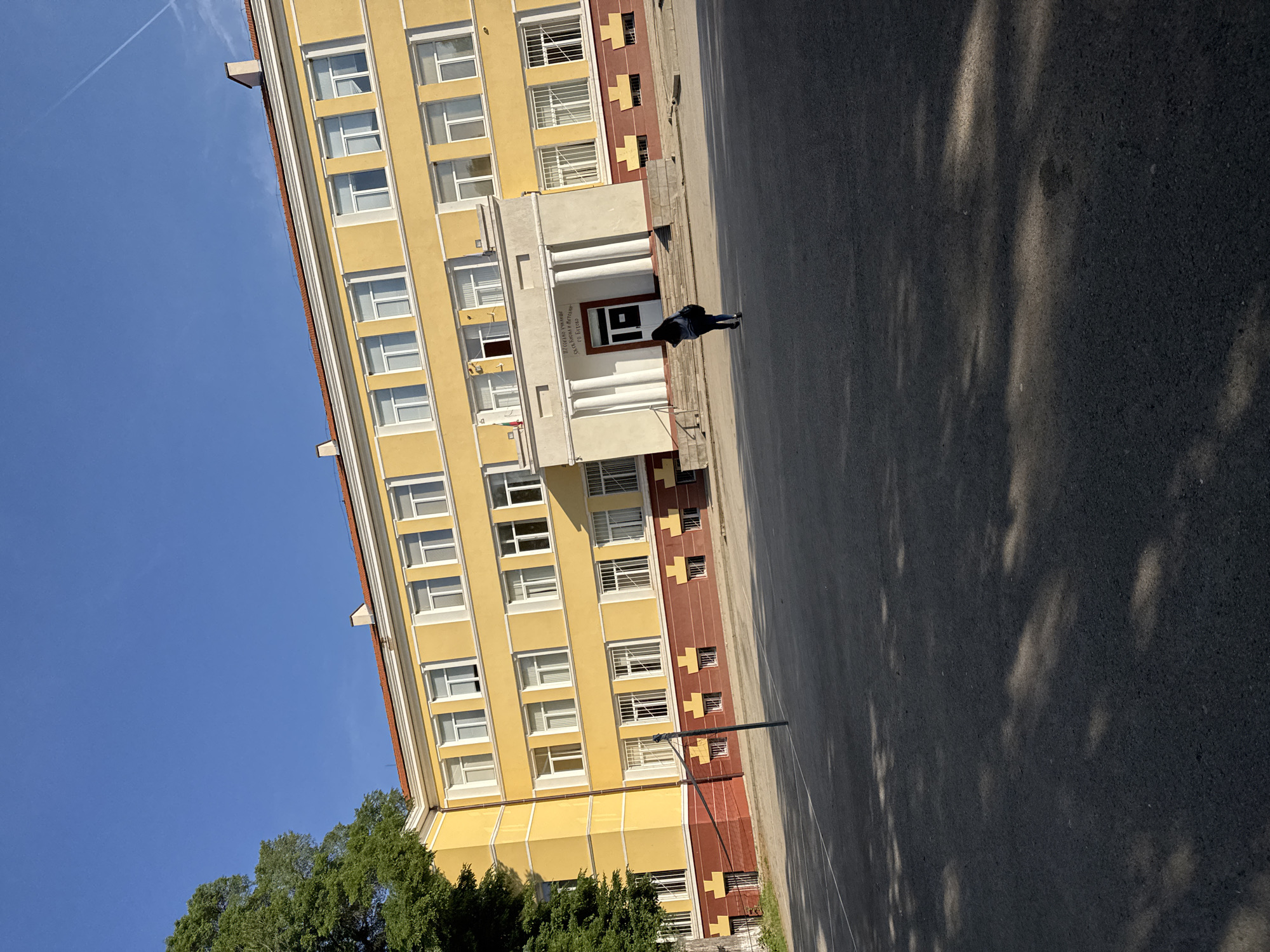 The image depicts a yellow, multi-story building with white-framed windows. A section of the building’s lower portion has a red and brown design with small square accents. The building’s entrance is marked by white pillars and a sign, bearing the text "ПРОФИЛИРАНА ГИМНАЗИЯ ПО ЕЛЕКТРОТЕХНИКА" and "Св. Арх. Михаил и Гаврил". A person wearing dark clothing walks in front of the building. The scene appears to be an exterior shot on a sunny day. The location is in Buhovo, Bulgaria. A portion of a tree’s green foliage is visible at the bottom left. The foreground is asphalt, with shadows from trees. There is a contrail in the clear blue sky. A lamppost stands on the street.