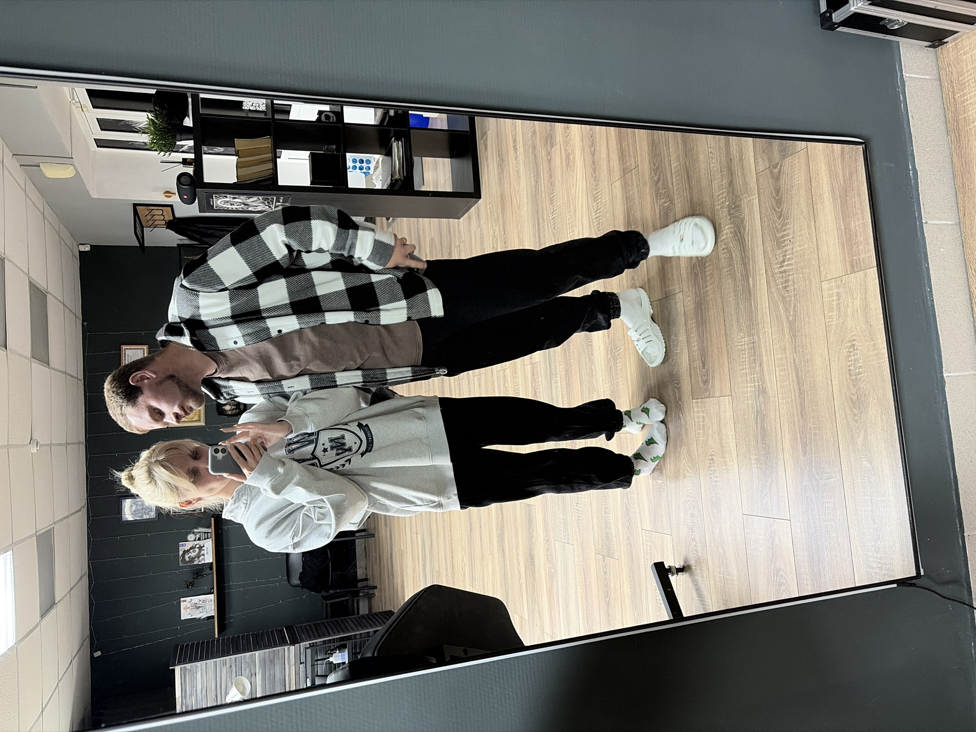 A selfie is taken in what appears to be a room with light-colored wood-patterned flooring and dark gray walls. The reflection in the mirror shows two people: a man with blonde hair and a woman with blonde hair pulled into a bun. The woman is holding a cell phone and taking the photo. The man is wearing a brown undershirt and a black and white plaid shirt, while the woman is wearing a light gray sweatshirt with a crest-like design on the front. The man is wearing black pants and white shoes, while the woman is wearing black pants and white socks with green patterns. In the background, a shelving unit filled with various items is visible. The room appears to be a retail or commercial space, potentially a tattoo parlor due to the decor on the wall. The location is possibly in Chaikovsky, Russia.