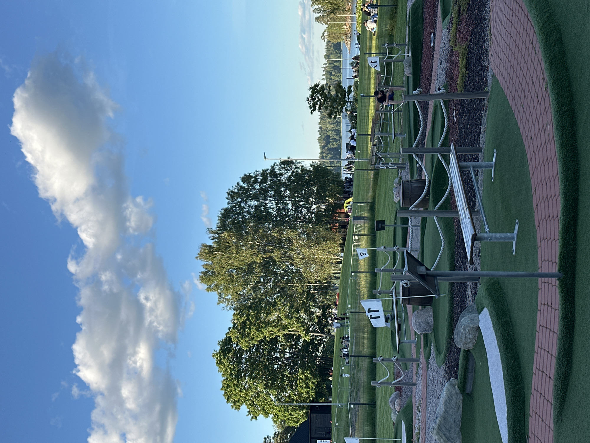 The image depicts an outdoor mini-golf course in a park-like setting. The foreground shows a portion of the mini-golf course with artificial turf, brick-like pathways, rocks, and wooden benches. Several flags, some with numbers or symbols, are planted throughout the course. In the middle ground, a group of people are gathered on a grassy hill, some sitting on blankets or at picnic tables. Beyond the hill, a body of water, likely a lake or bay, is visible with trees and a distant shoreline. The sky is blue with scattered white clouds. The overall scene suggests a recreational area on a pleasant day.