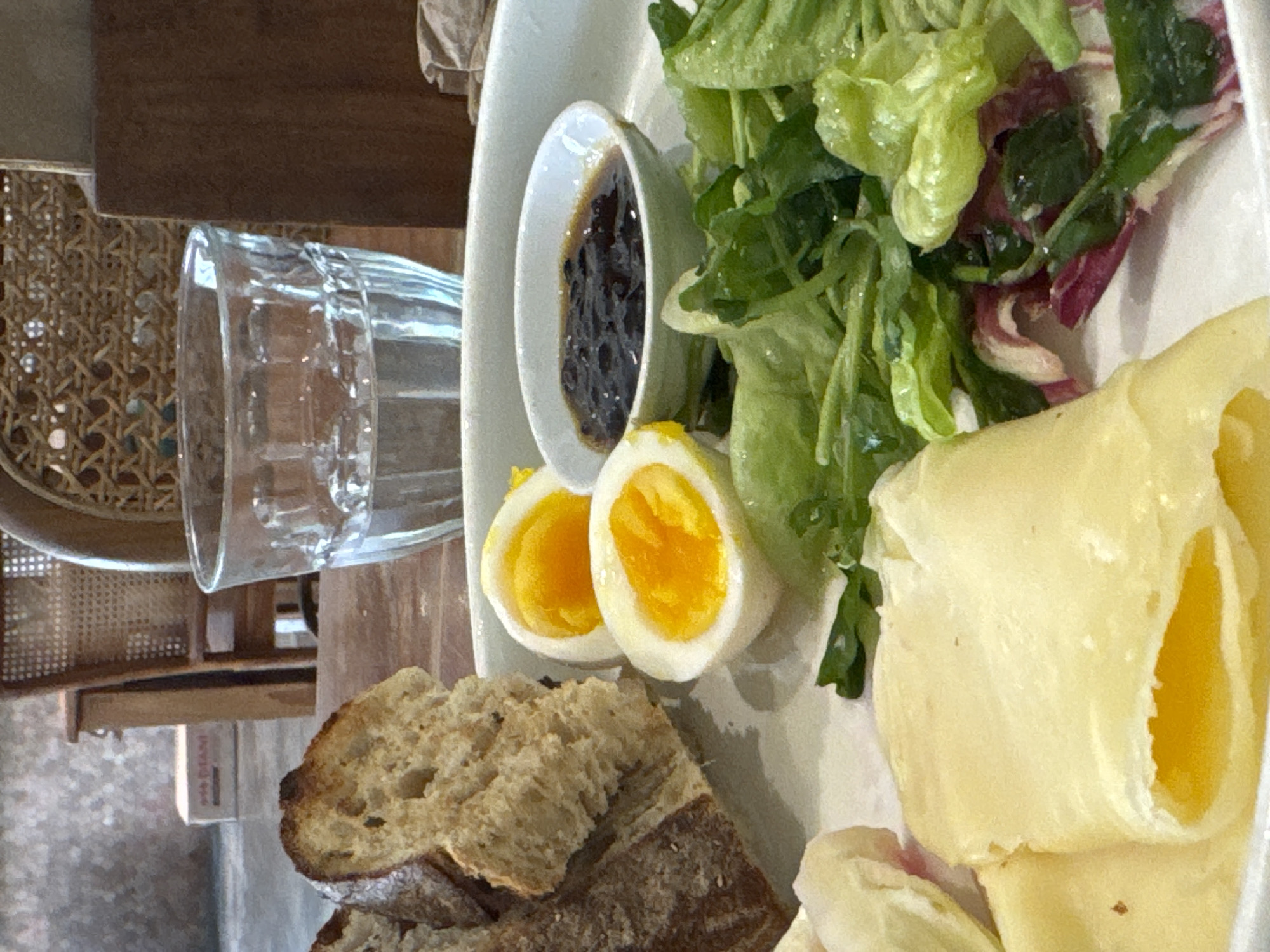 The image shows a plate with a salad, boiled eggs, bread, and a small bowl of sauce. A glass of water is next to the plate on a wooden table. A wicker chair is visible in the background. The image was taken in Ungasan, Indonesia.