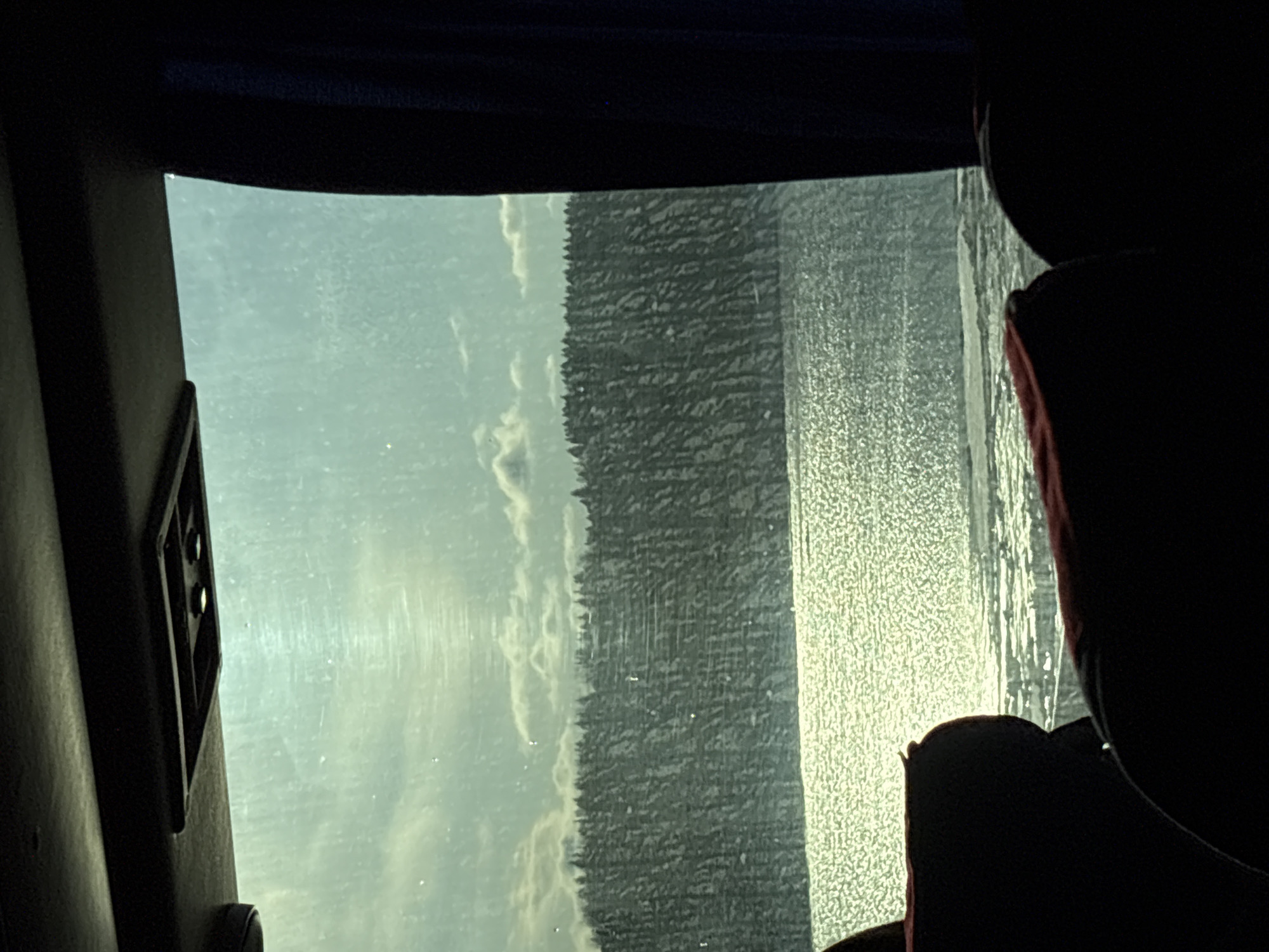 The image appears to be taken from inside a vehicle, possibly a bus or train, looking out a window. The exterior scene includes a sky with clouds, a dense forest, and possibly a body of water or snow-covered field reflecting sunlight. There's a seat visible within the vehicle. The location is potentially Usolye, Russia.