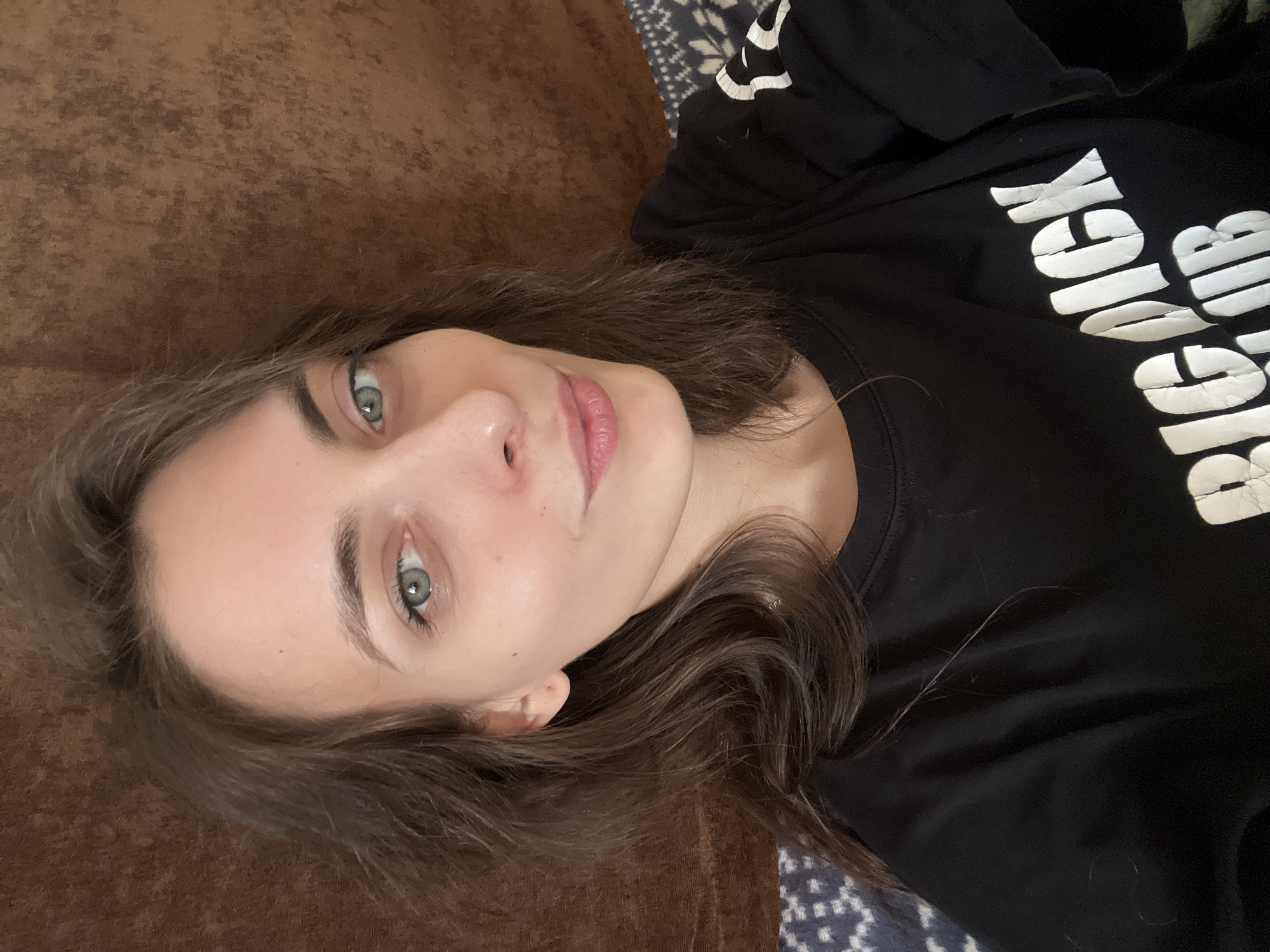A young woman with fair skin, brown hair, and blue eyes lies on a brown, textured surface, possibly a couch. She wears a black t-shirt with white lettering, part of which is visible and reads "Bigder." She looks towards the camera with a neutral expression. A patterned blue and white textile is partially visible underneath her. The location is unknown and there is no information to determine it is Krasnoyarsk, Russia.