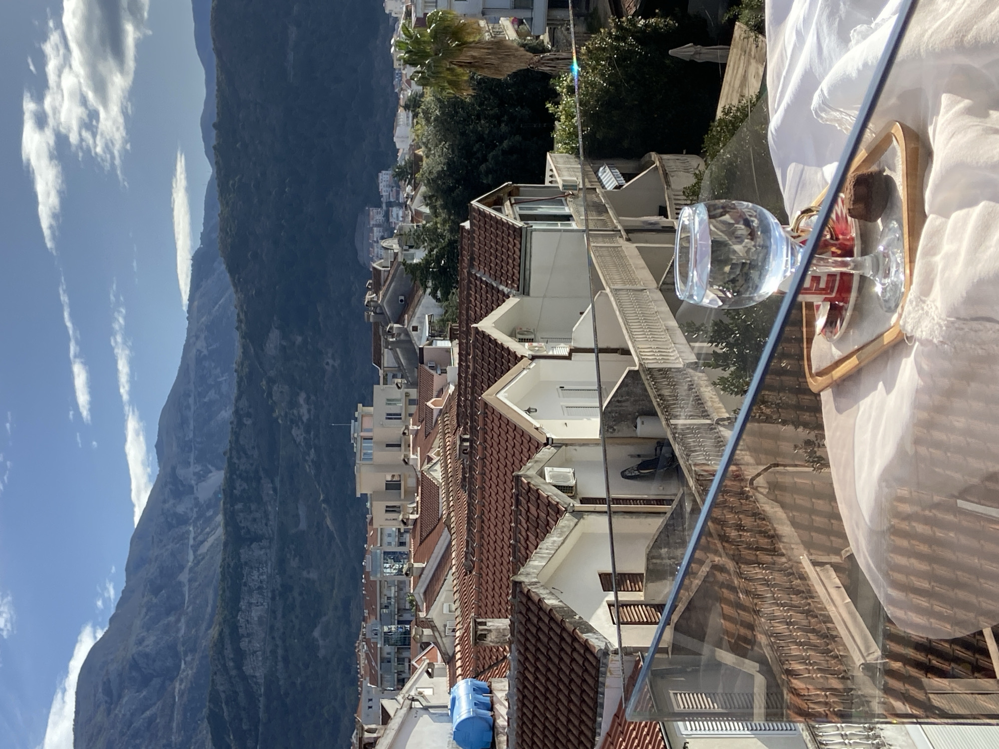 A view of a city with reddish-brown tiled roofs and white walls is seen, with a large mountain in the background under a blue sky with scattered clouds. A glass of water, a saucer, and a small dark object are on a wooden tray, sitting on a white surface. The view appears to be from a balcony or window, as the roof and walls of the adjacent buildings are visible. The scene likely depicts Budva, Montenegro, given the architecture and landscape. No people are visible.