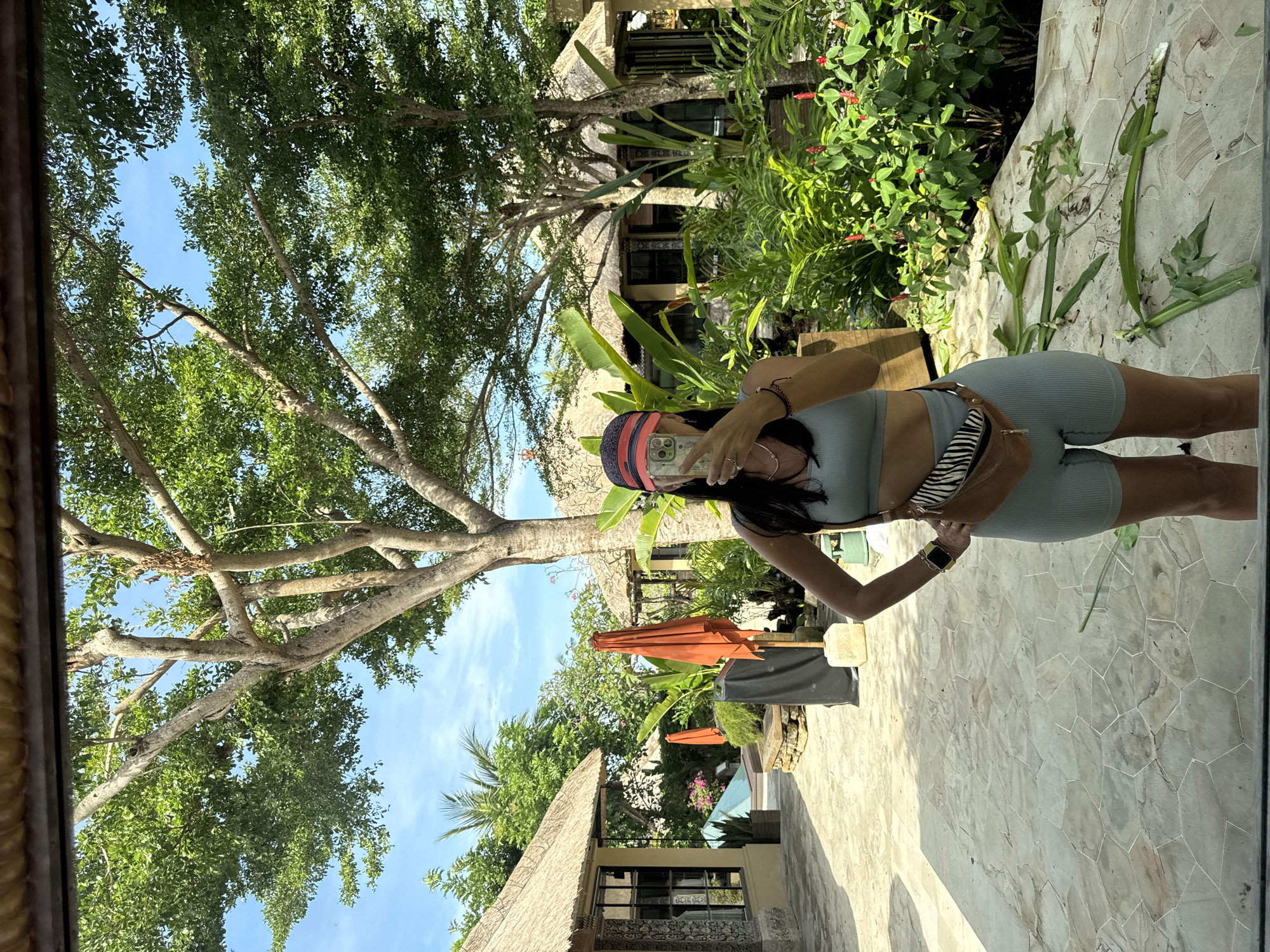 The image is a selfie taken in Kuta Selatan, Indonesia. A woman stands facing a mirror, holding a phone to take the photo. She is wearing a matching light blue athletic set and a baseball cap with a striped pattern. A zebra-print purse is slung over her shoulder. Behind her, the scene transitions from an outdoor patio with tiled flooring to a lush tropical garden. The garden features a variety of plants and flowers, and there are thatched-roof structures visible. An orange parasol adds a splash of color. The sky is blue with scattered clouds, and a large tree with dense foliage dominates the left side of the frame. The overall impression is that the photo was taken in a resort or villa-like setting.