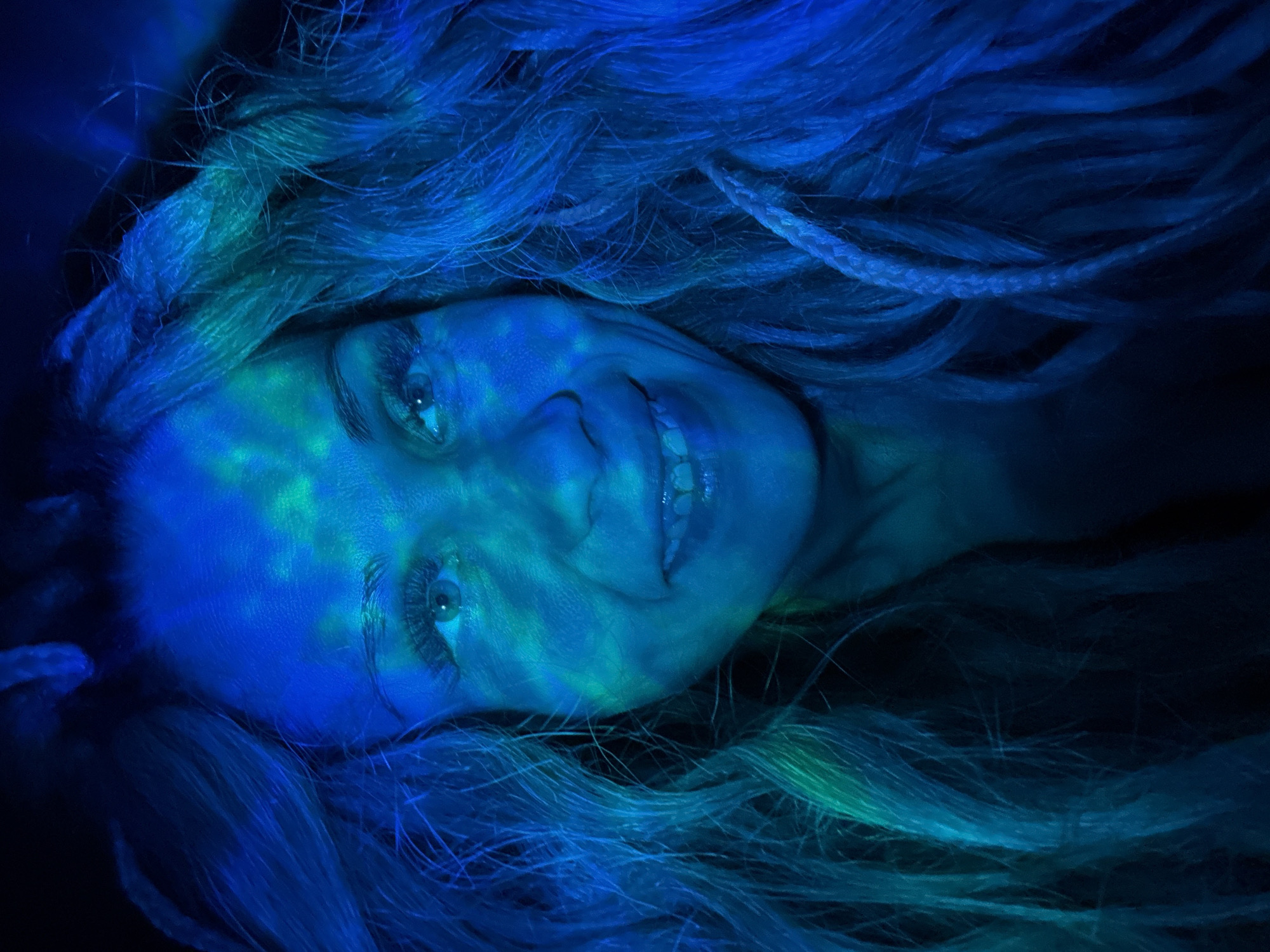 A woman lies on her back, her face partially illuminated with a blue and green light pattern. She has light-colored hair that surrounds her face, some of which appears to be braided. Her eyes are open, and she is smiling slightly, revealing her teeth. The background is dark and out of focus. There are no visible indicators that the scene is in Rusanovo, Russia.