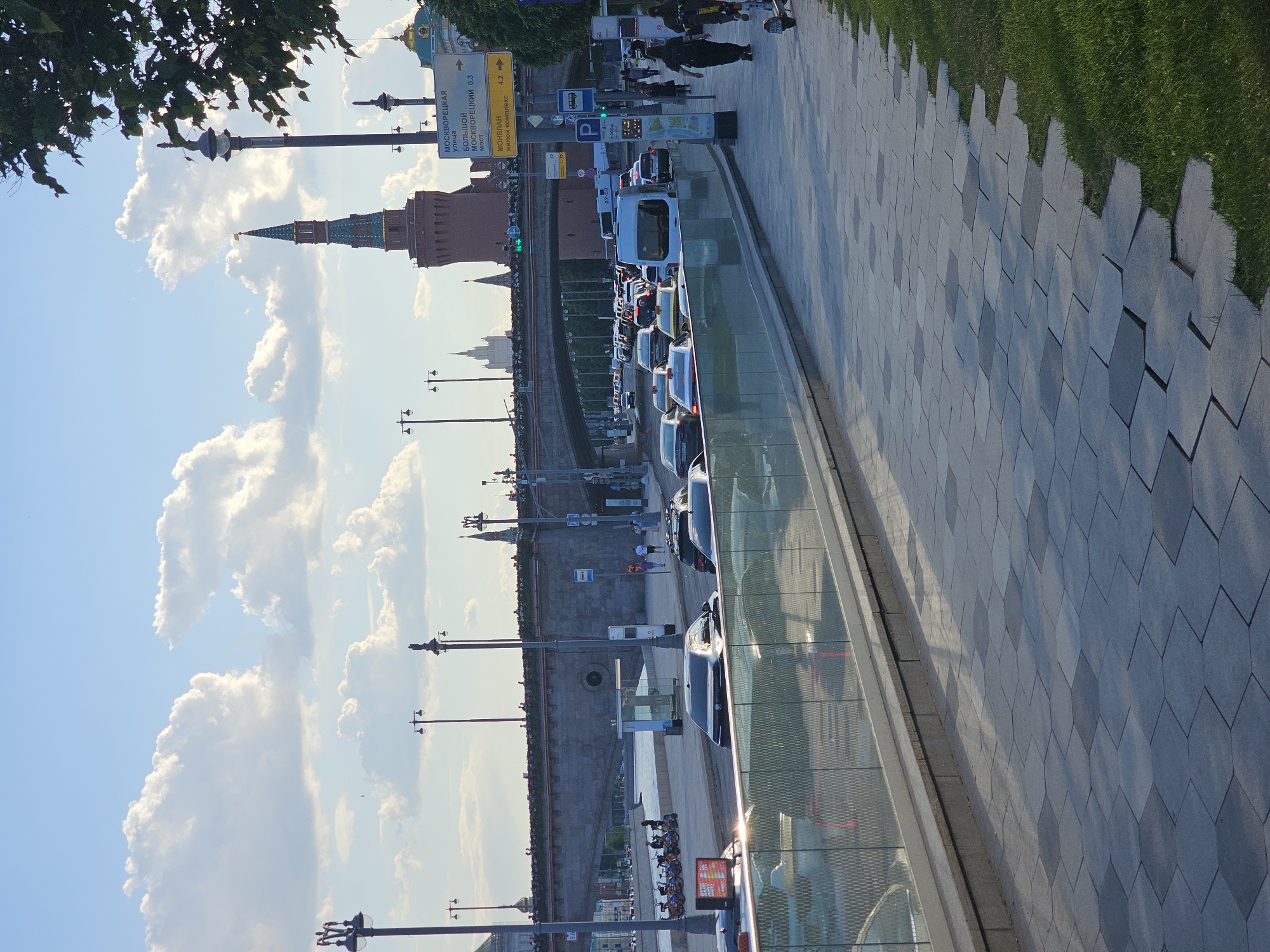 A bright, sunny cityscape captures a street view with the Moscow Kremlin in the background. In the foreground, a wide pedestrian walkway is paved with geometric, possibly hexagonal, grey tiles, bordered by a strip of neatly maintained green grass on the far right. A modern glass barrier runs alongside the paved area, separating it from the lower level of the street.
The midground features a multi-lane road with moderate traffic, including several dark-colored cars and a prominent white van. Pedestrians are visible walking along the pavement further down the street, some near a bus stop sign and other street signs. Tall black streetlights line the road and the adjacent embankment.
In the background, the distinctive red-brick walls and towers of the Moscow Kremlin are clearly visible under a vast blue sky dotted with fluffy white clouds. A prominent tower with a green-tiled spire and a small yellow element at its peak stands out.
Visible text on yellow street signs includes "МОСКВОРЕЦКАЯ УЛИЦА" (Moskvoretskaya Street) and "БОЛЬШОЙ МОСКВОРЕЦКИЙ МОСТ" (Bolshoy Moskvoretsky Bridge) with a distance of "0.3". Another sign points left, "<- 4.2", to "МОНЕПАН ЖИЛОЙ КОМПЛЕКС" (Monetpan Residential Complex). Blue signs indicate a bus stop and parking ("P"). The time of day appears to be late morning or early afternoon, given the bright sunlight and clear shadows.