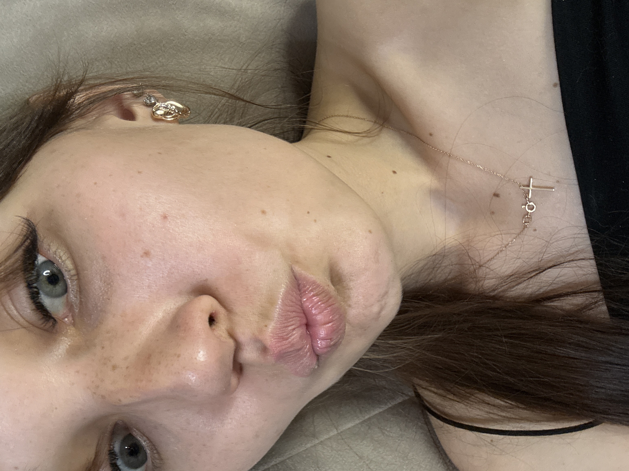 This is a close-up, over-the-shoulder portrait of a young woman with light skin, freckles, and blue-grey eyes, wearing a black tank top and a rose gold necklace with a cross pendant. She has on makeup, including eyeliner and mascara, and her lips are slightly parted in a pout. She is also wearing a gold heart-shaped earring with small diamonds on her right earlobe. Her dark brown hair is visible on the left side of the frame. The background appears to be a textured, light-colored surface, possibly upholstery. The image does not contain any elements that would suggest a specific location in Novokuznetsk, Russia.