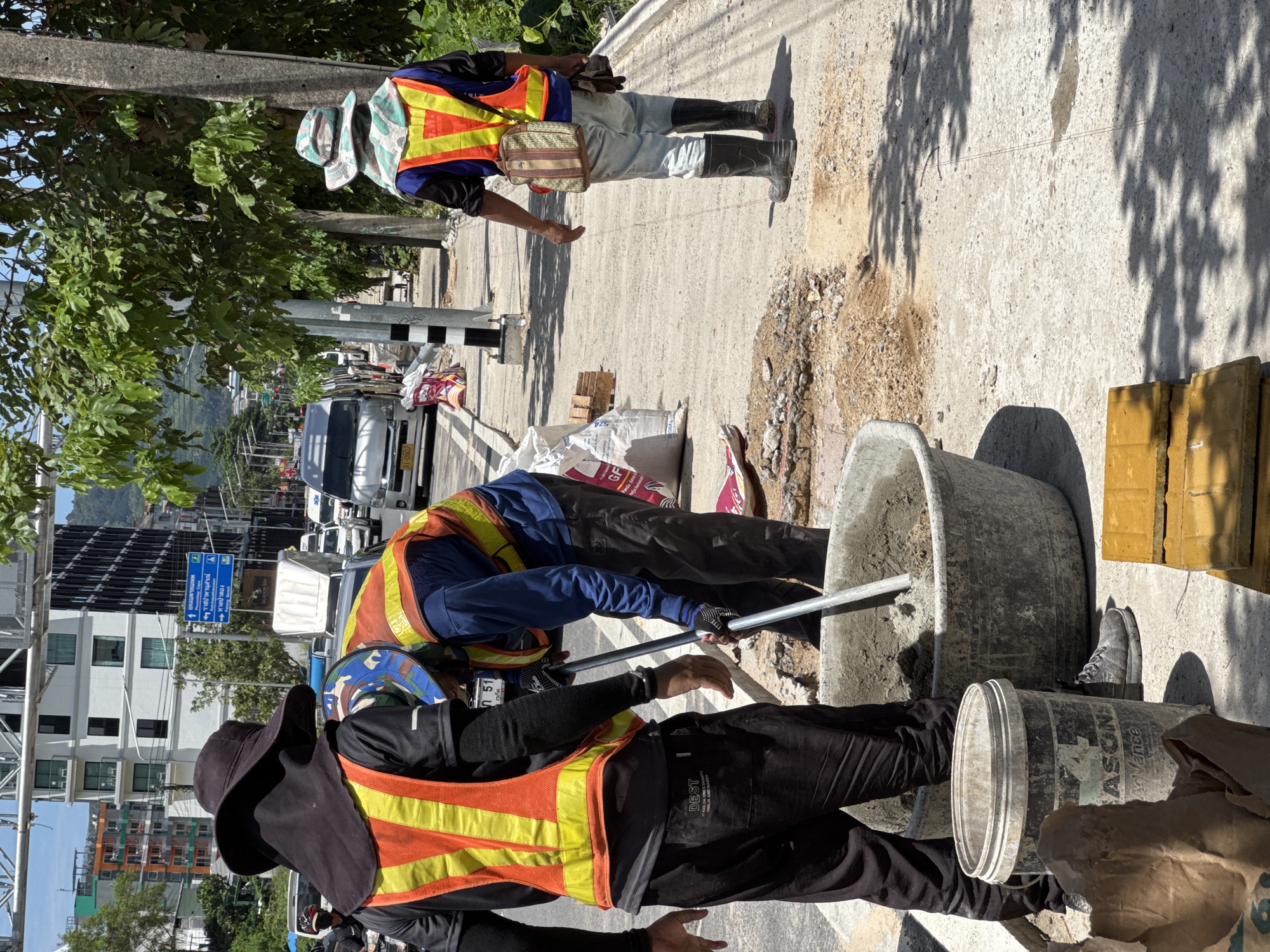 The image depicts a street scene in Patong, Thailand, where construction work is underway on a sidewalk. Two construction workers, dressed in high-visibility vests and protective headwear, are actively engaged in mixing cement in a large grey basin. One worker is operating a long-handled tool within the basin, while the other stands nearby, observing. Various construction materials, including bags of cement, loose sand and gravel, and yellow paving stones, are scattered around the work area. In the background, a street with passing vehicles, buildings, and lush green trees indicates an urban environment. The overall impression is of ongoing infrastructure maintenance or development in a tropical setting.