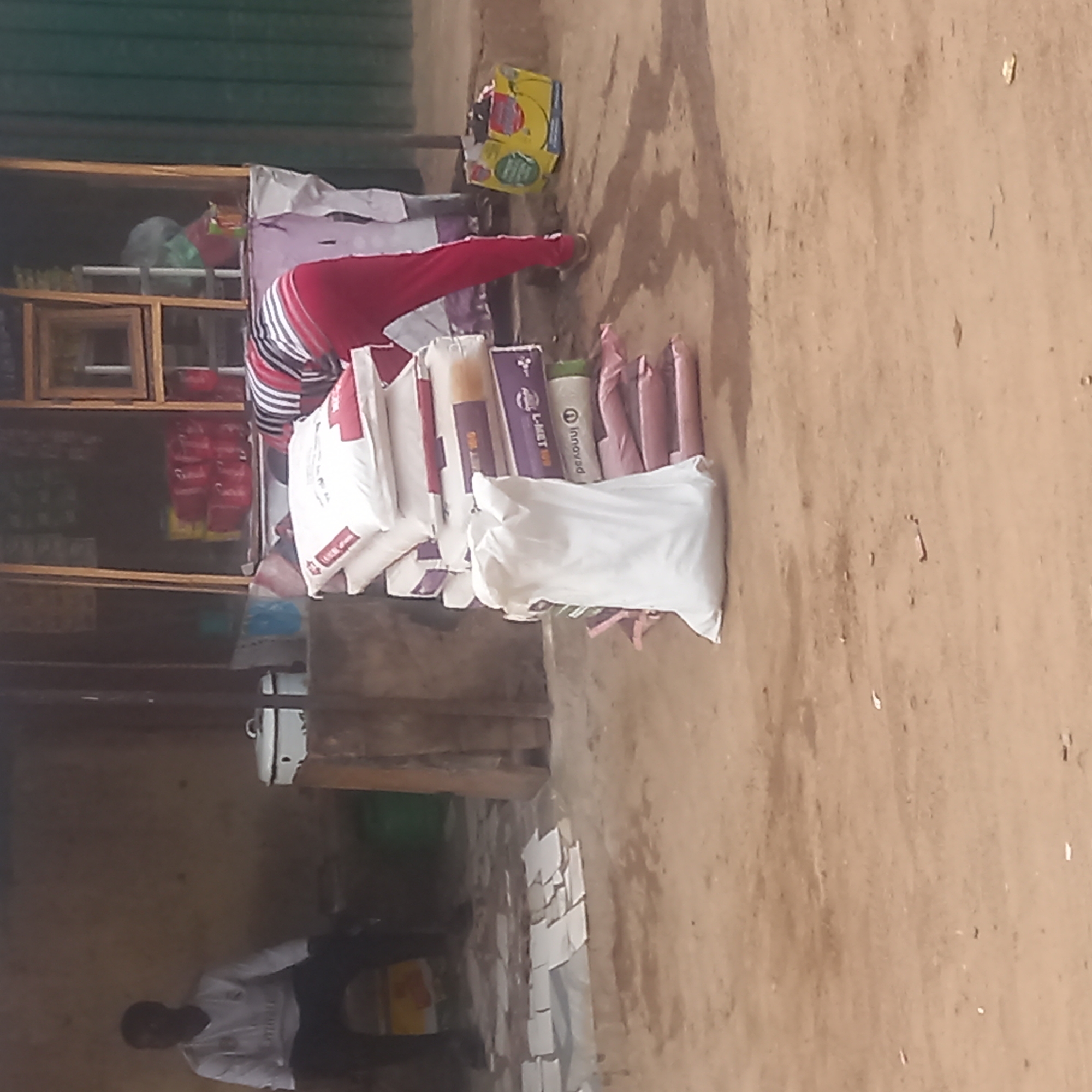 The image depicts a scene in Dadi Riba, Nigeria. A person wearing red pants is standing near a wooden shelf loaded with various goods. Several bags, possibly containing products for sale, are arranged on the ground in front of the shelf. A man in a white shirt and dark pants is visible in the lower left corner of the image. The ground appears to be a dirt surface. The environment suggests a small retail or commercial space.