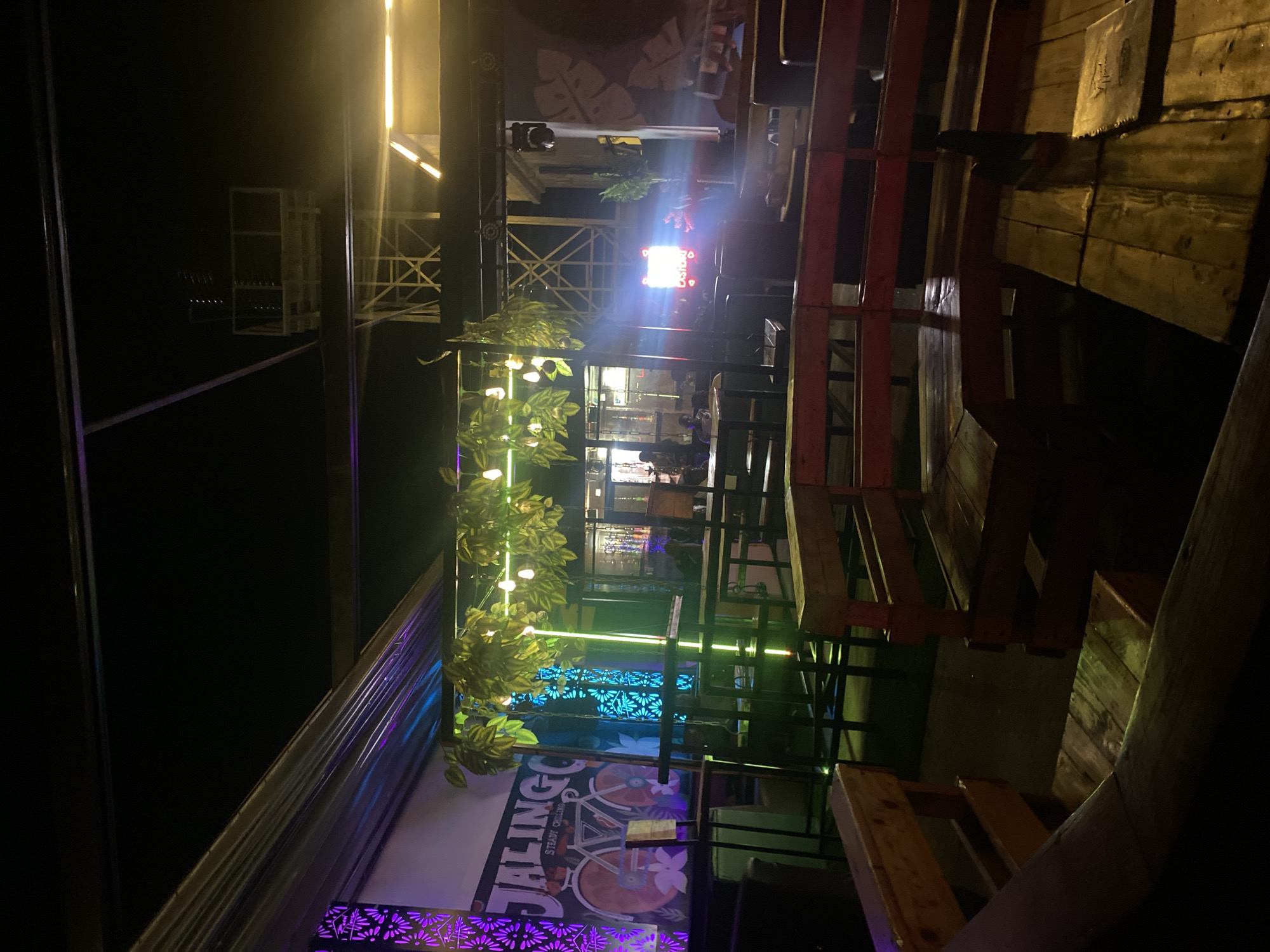 The image depicts a dimly lit outdoor seating area of a establishment, possibly a bar or restaurant, at night. The foreground is dominated by wooden benches and tables. In the background, there are neon lights, decorative plants, and structures that suggest a lively atmosphere, possibly a music venue or a social gathering spot. The overall ambiance is dark with pops of vibrant color from the lighting. The location in the city of Howayi, Nigeria is inferred from the user's prompt and is not visually evident in the image itself. There are a few indistinct figures visible in the background, implying the presence of patrons.