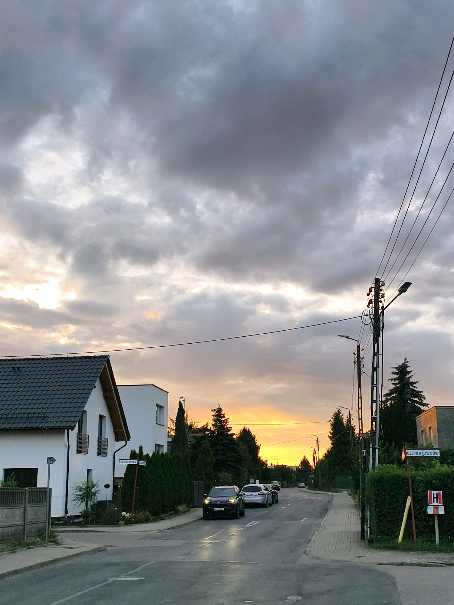 The image shows a street scene in Tczew, Poland, at dusk. The sky is cloudy with streaks of orange and yellow from the setting sun. A modern white house with a dark tiled roof is on the left side of the street. Several cars are driving on the road, and there are streetlights and utility poles visible. A street sign for "UL. CHLOPSKA" is mounted on a pole near the house. Further down the street, another street sign reads "UL. PORTOWCOW" and displays a red "H" symbol, indicating a hospital. Trees and hedges line the sides of the road.