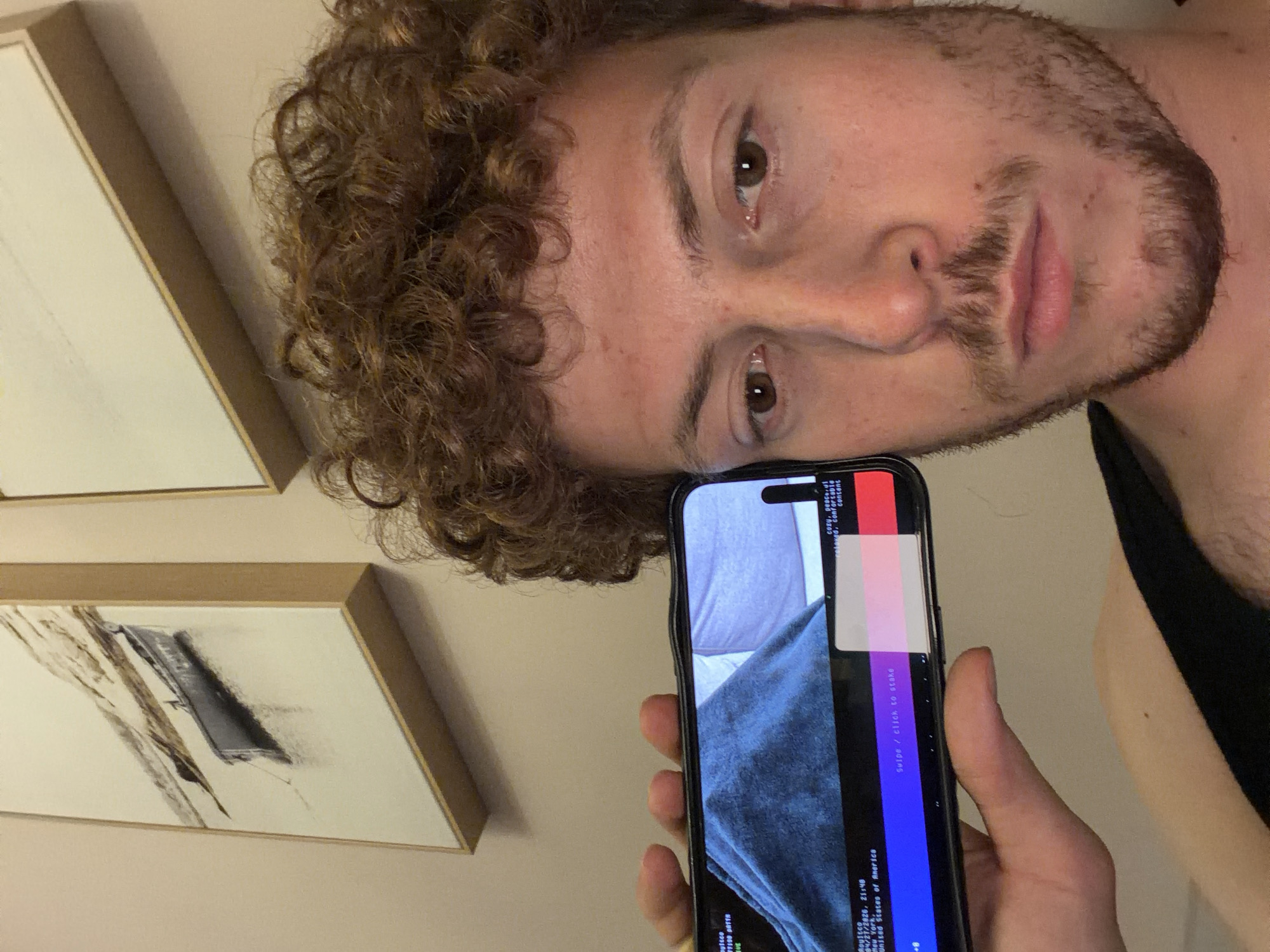 A young man with curly brown hair and light brown stubble/beard is depicted in a close-up, rotated shot, looking directly at the viewer with a neutral expression. He is wearing a black sleeveless top and holds a smartphone in his left hand. Behind him, two framed abstract artworks hang on a light-colored wall. The phone screen displays a partially glitched image featuring colorful bars (red, white, pink, purple, blue, black) overlaying what appears to be grey-blue fabric.
Visible text on the phone screen includes: "multco", "#7190 PATTH", "Roustco", "9/27/2026, 21:40", "New York, United States of America", "Swipe / click to stake", "cozy, peace all", "and, comfortable", and "consensus". The time stamp "21:40" suggests the photo was taken at night. The embedded text also indicates the location is New York, United States of America.