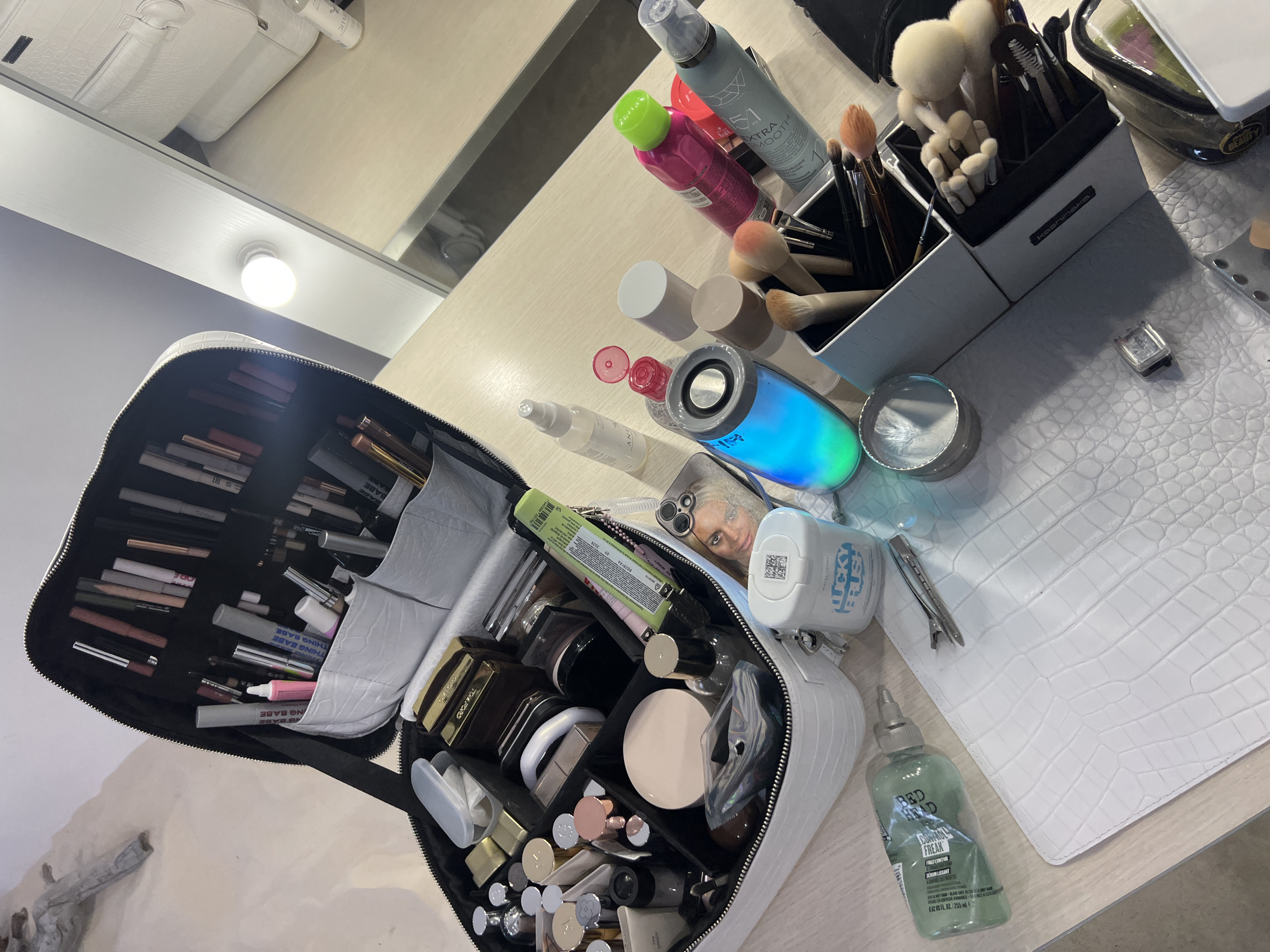 An overhead, slightly angled shot reveals a collection of beauty and grooming products spread across a light-colored wooden or laminate countertop. The scene appears to be a vanity or dressing area, with a mirror reflecting a bright light bulb in the upper background.
The primary subject is a large, open white makeup bag with a crocodile-embossed texture, brimming with numerous cosmetic items. Inside, various lipsticks, foundations, compact powders, and brushes are neatly arranged. Text like "THING BABE" and "TOM FORD" is visible on some of the products within the bag.
To the right of the bag, an assortment of other items is displayed on the counter. These include two white organizers filled with various makeup brushes, one of which is labeled "kashinaka". A clear, cylindrical speaker emits vibrant blue, green, and purple light. Nearby, several spray bottles are present, including a grey one with "XTRA SMOOTH" text and a bright pink one. A clear bottle of "BED HEAD CONTROL FREAK FRIZZ CONTROL SERUM LISSANT" (255 FL. OZ.) stands towards the bottom right. A white container marked "LUCKY BRUSH" and a small, square silver-toned watch are also on the counter. A phone case featuring a printed image of a woman's face is partially visible near the open makeup bag. The countertop itself is partially covered by a white, crocodile-textured mat or case.
The arrangement suggests preparation for a makeup session or styling, or perhaps the organized toolkit of a beauty professional. The lighting is artificial, originating from the visible bulb in the mirror.