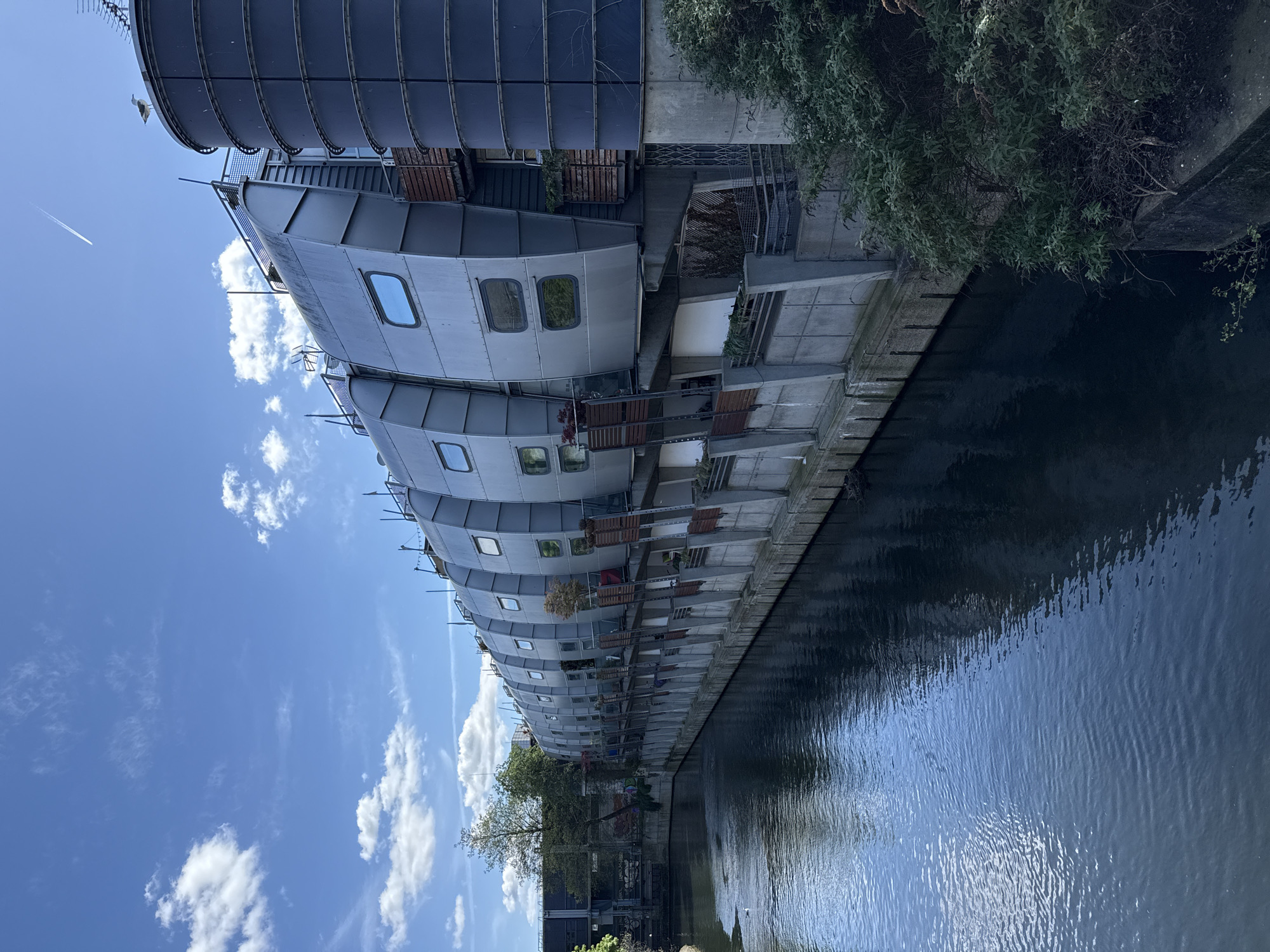 The image shows a row of modern, curved buildings situated along a canal in the city of London, United Kingdom. The buildings appear to be apartments or houseboats, characterized by their distinctive rounded shapes and metallic exteriors. They feature rectangular windows and small balconies. The canal water is relatively still, reflecting the buildings and the sky. The sky is blue with scattered white clouds. Vegetation, including trees and bushes, is visible along the canal bank. The image appears to be taken during daylight hours, likely in the late morning or early afternoon. A contrail from a plane can be seen streaking across the sky.
