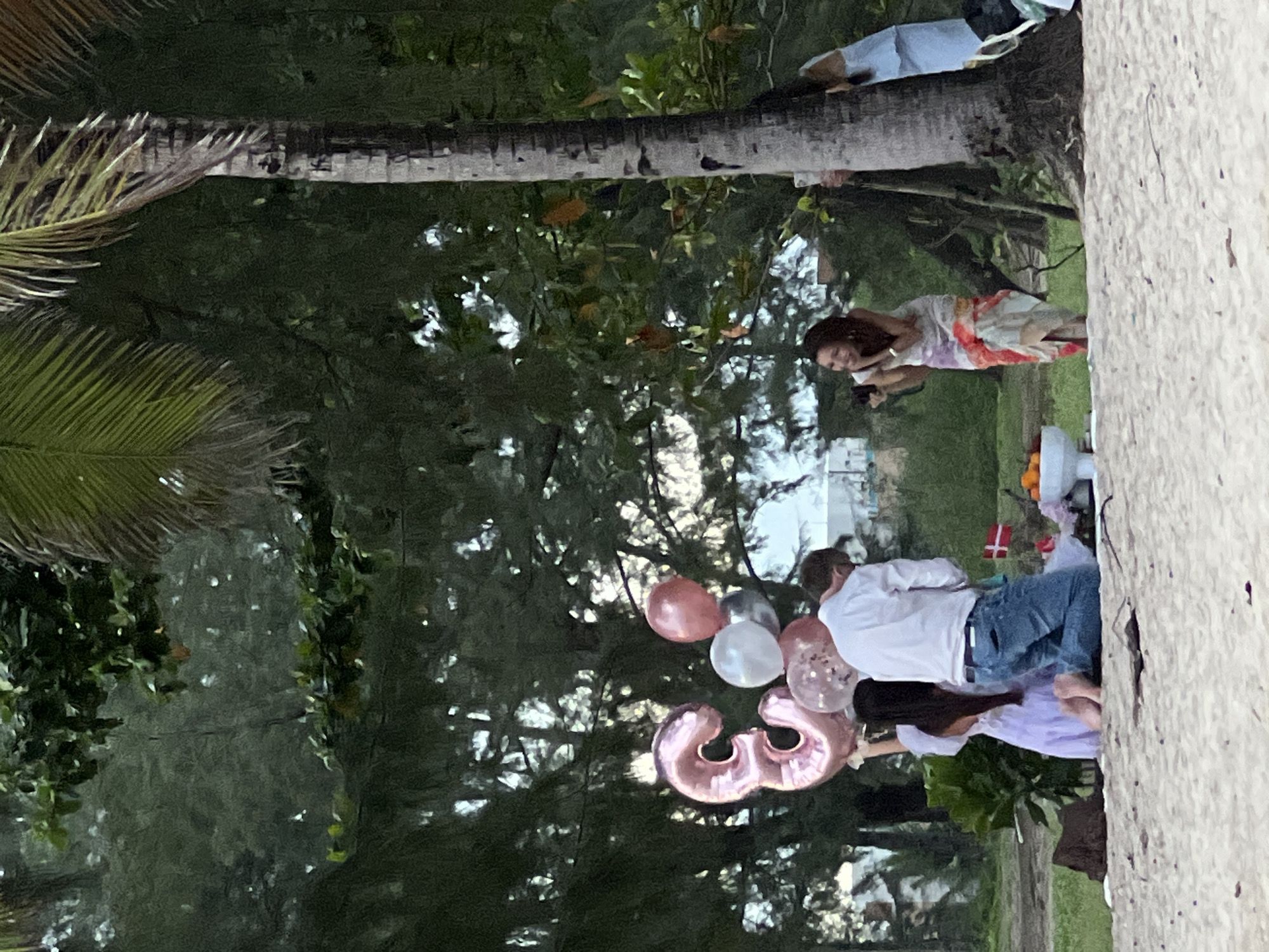 On a sandy beach, a group of people are gathered near a palm tree. In the foreground, a large, pink, foil balloon shaped like the number "3" is held by a person dressed in a light purple garment. Next to them, a person in a white shirt and blue jeans is kneeling. A collection of balloons, including pink, white, and silver, are also present. In the background, a woman in a colorful dress is standing and holding a camera. The environment is lush with trees and foliage.