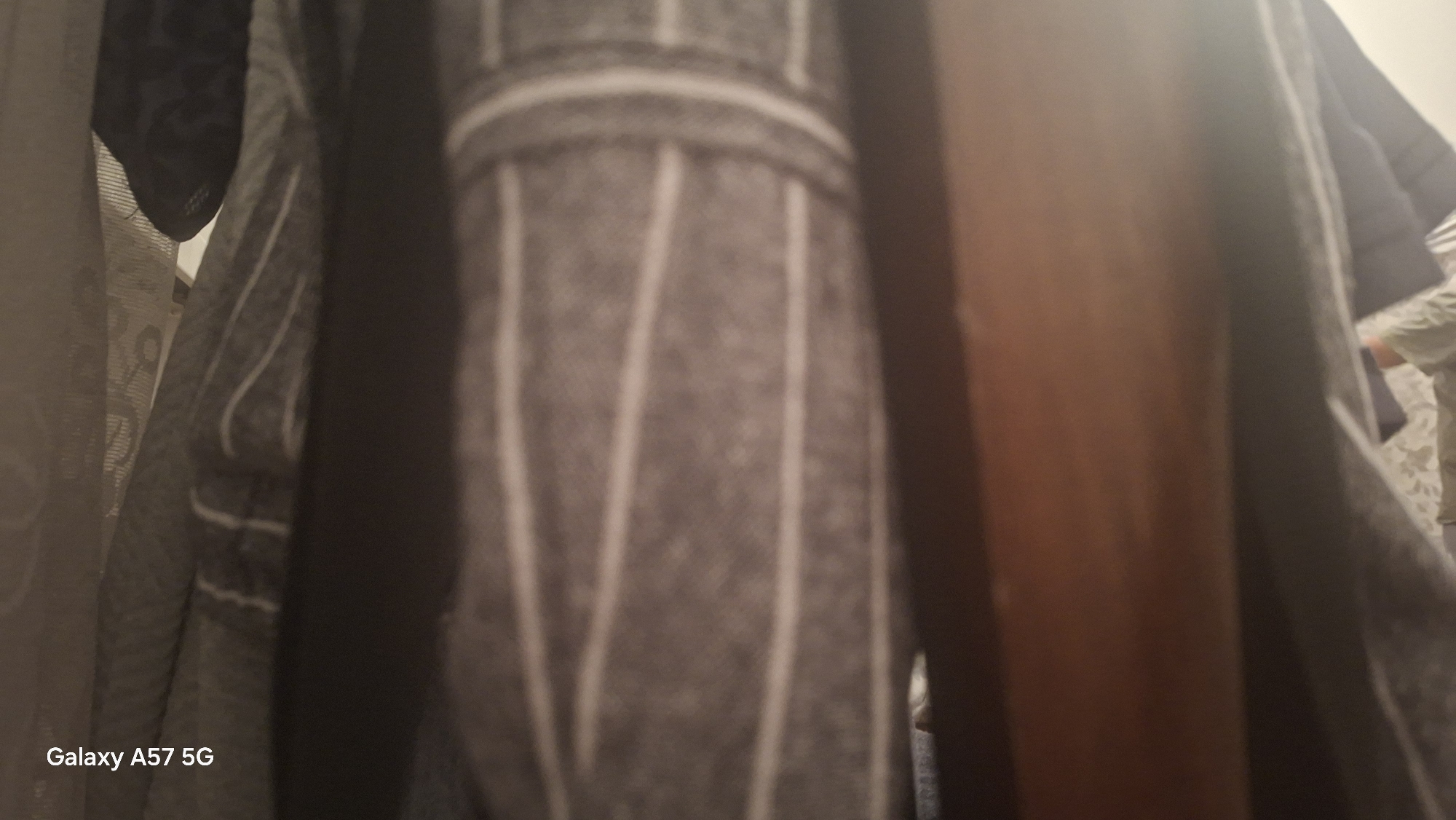 The image shows a close-up view of clothing hanging in a closet or wardrobe. The dominant element is a gray fabric with vertical white stripes, possibly a shirt or jacket. To the left, another garment made of a lighter, textured fabric is visible. A dark, vertical object, likely part of the closet structure, is positioned in the center-right of the frame. In the background, on the far right, a person's arm and shoulder are visible, wearing a light-colored, patterned garment. The overall lighting is dim, suggesting an indoor environment. There is no indication of Agarak, Armenia, or any other specific location in the image.