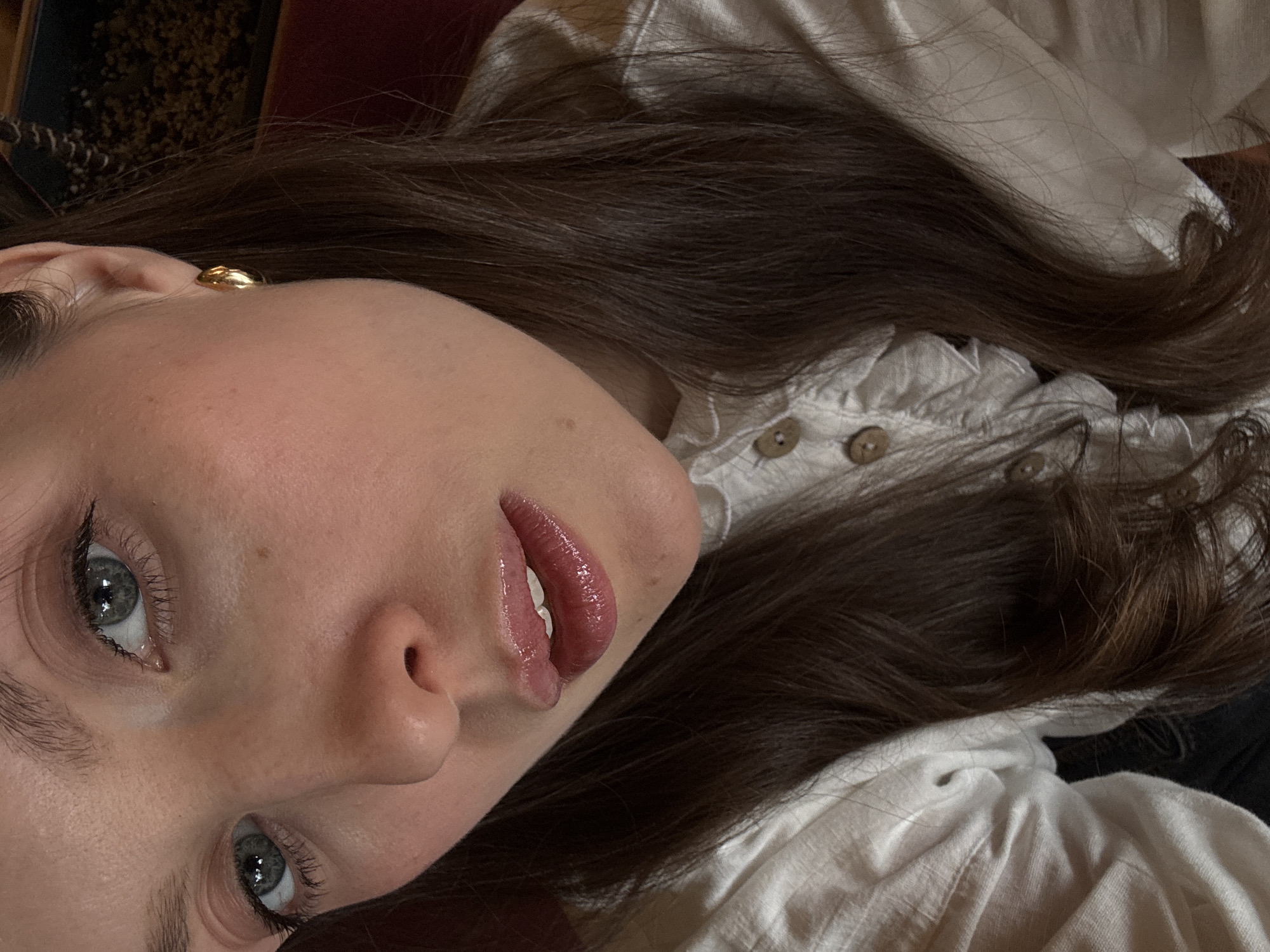 A close-up, tilted, indoor photograph depicts a person with fair skin, brown hair, and light-colored eyes. The person's face is angled to the left. Their lips are glossy. The person is wearing a light-colored shirt with small, round buttons visible near the collar. A gold stud earring is visible in the person's left ear. No location is discernible from the image.